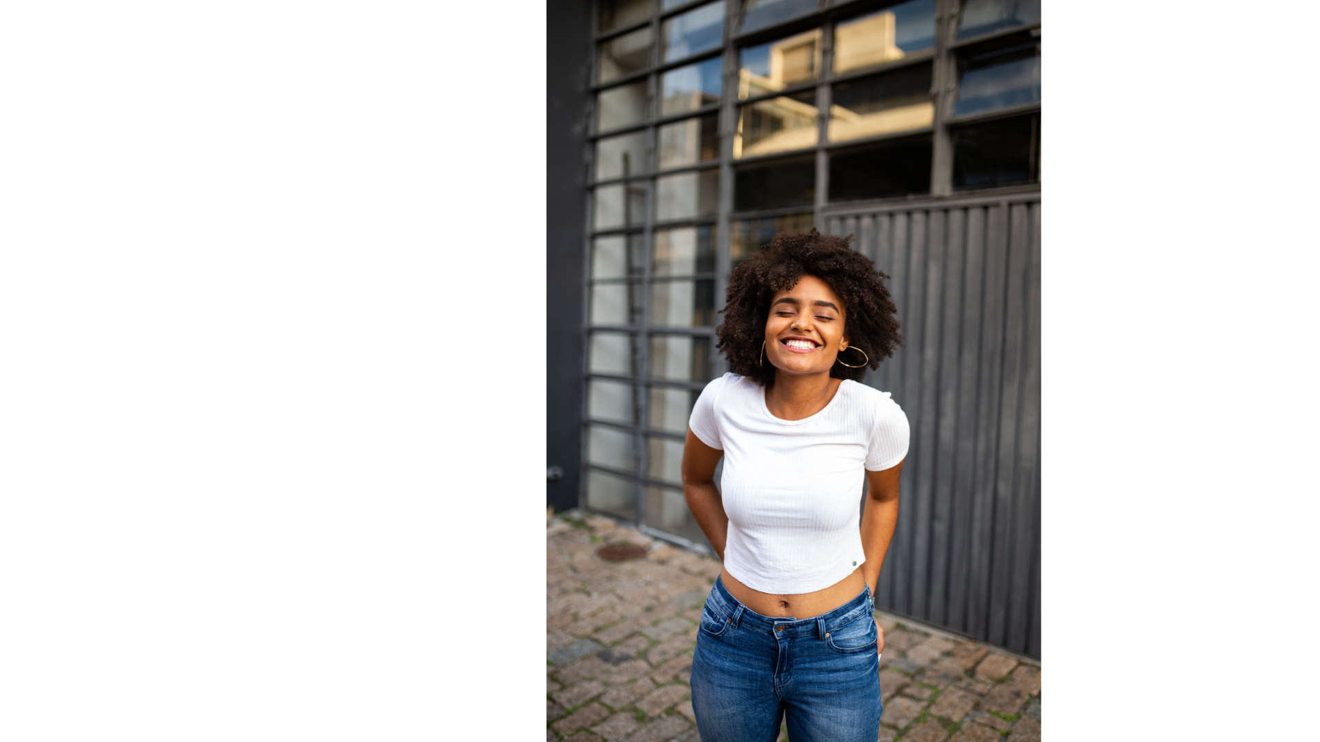 A young woman with curly hair smiling with eyes closed, wearing a white crop top and blue jeans, standing outdoors in front of a modern building with large windows and metal grid framework.