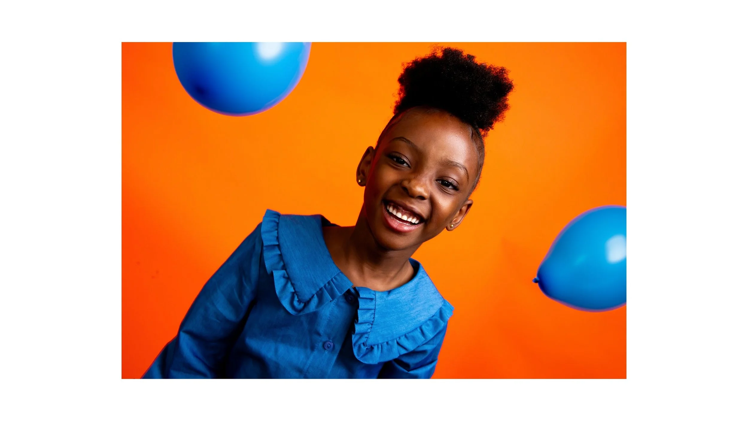 Smiling young girl with curly hair in a blue shirt, standing against an orange background, with blue balloons floating around her.