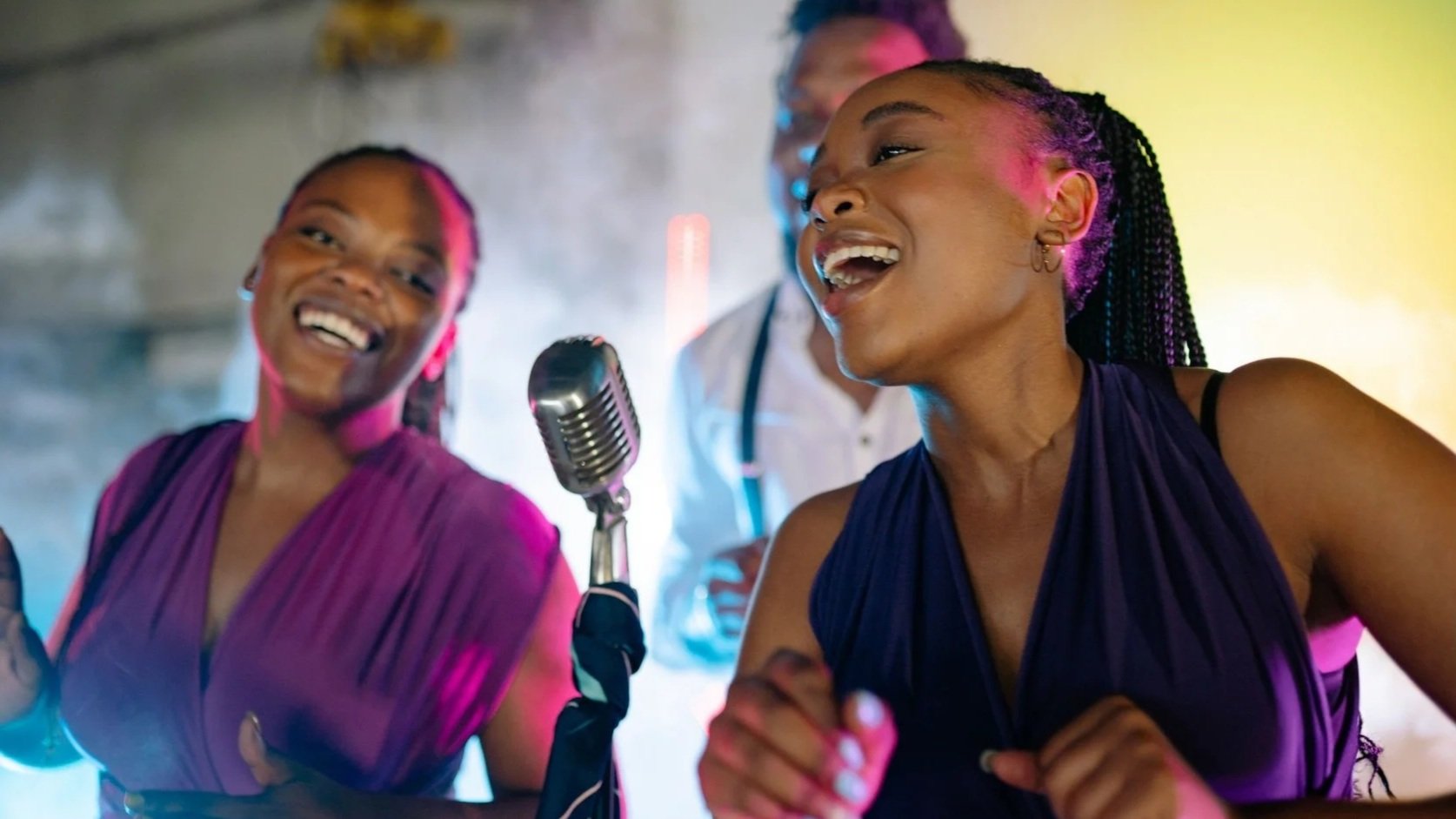 Two women with braided hair singing and laughing in front of a vintage microphone, with colorful lighting in the background.