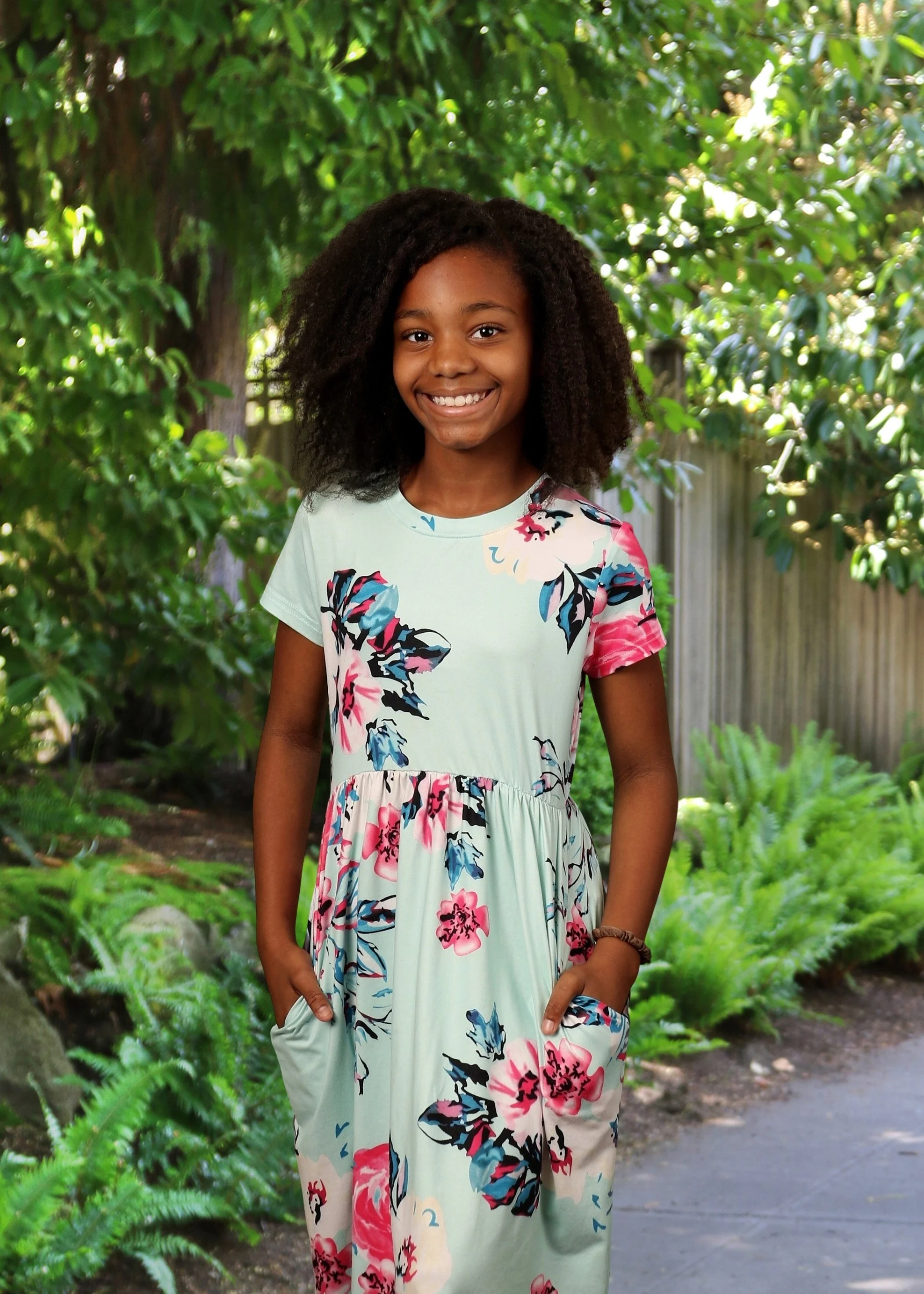 A young girl with dark curly hair standing outdoors in a garden with green foliage, smiling, wearing a floral dress with pink, blue, and black flowers.
