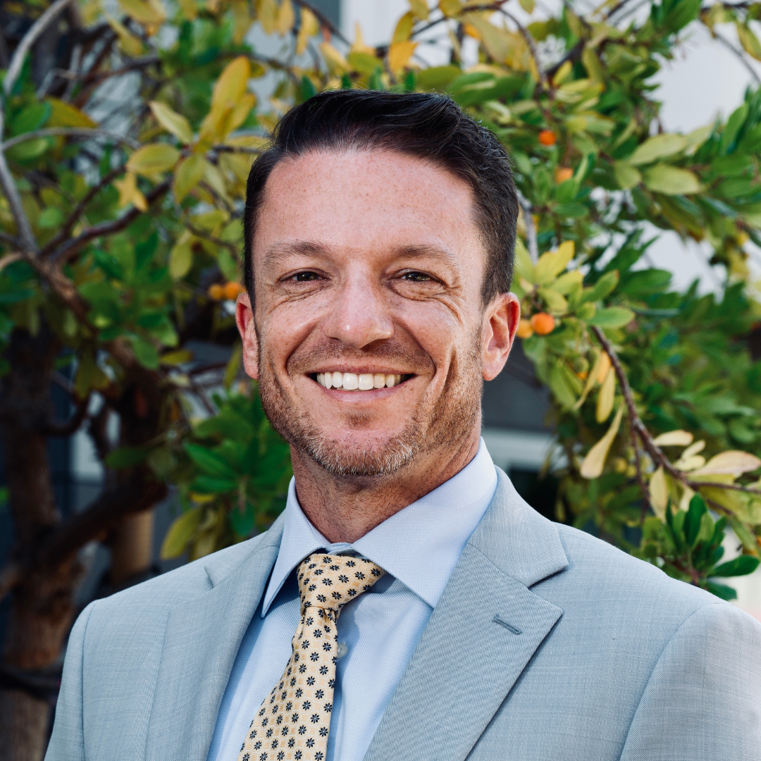Portrait of a smiling man in a light gray suit and patterned tie, standing outdoors in front of green foliage and a tree.