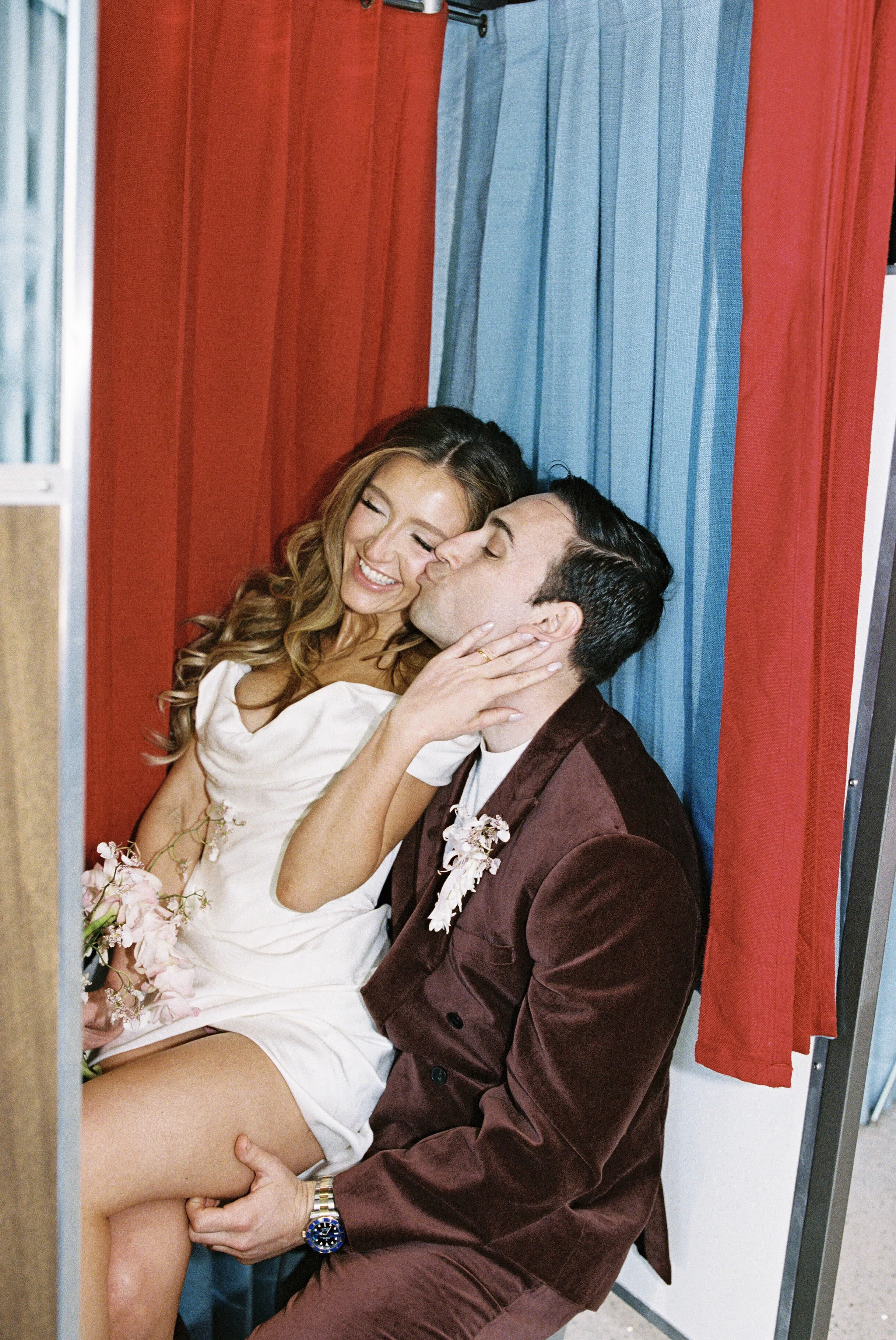 A woman and a man are sitting close together against a red and blue curtain. The woman has long, wavy hair, is smiling, and is wearing a white dress holding pink flowers. The man has dark hair, is wearing a maroon jacket with a white shirt, and is ki