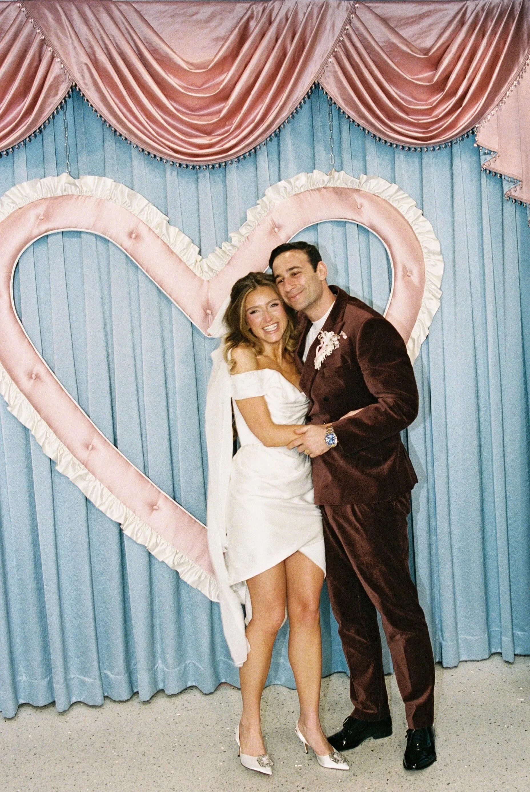 A smiling woman in a white dress and a man in a dark suit are embracing in front of a pink and blue heart-shaped backdrop.