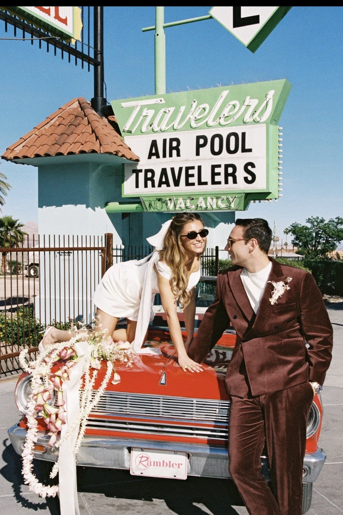 A couple dressed in wedding attire leaning on a vintage red car decorated with flowers, parked in front of a travel agency sign that advertises air pool travelers, with a clear blue sky overhead.