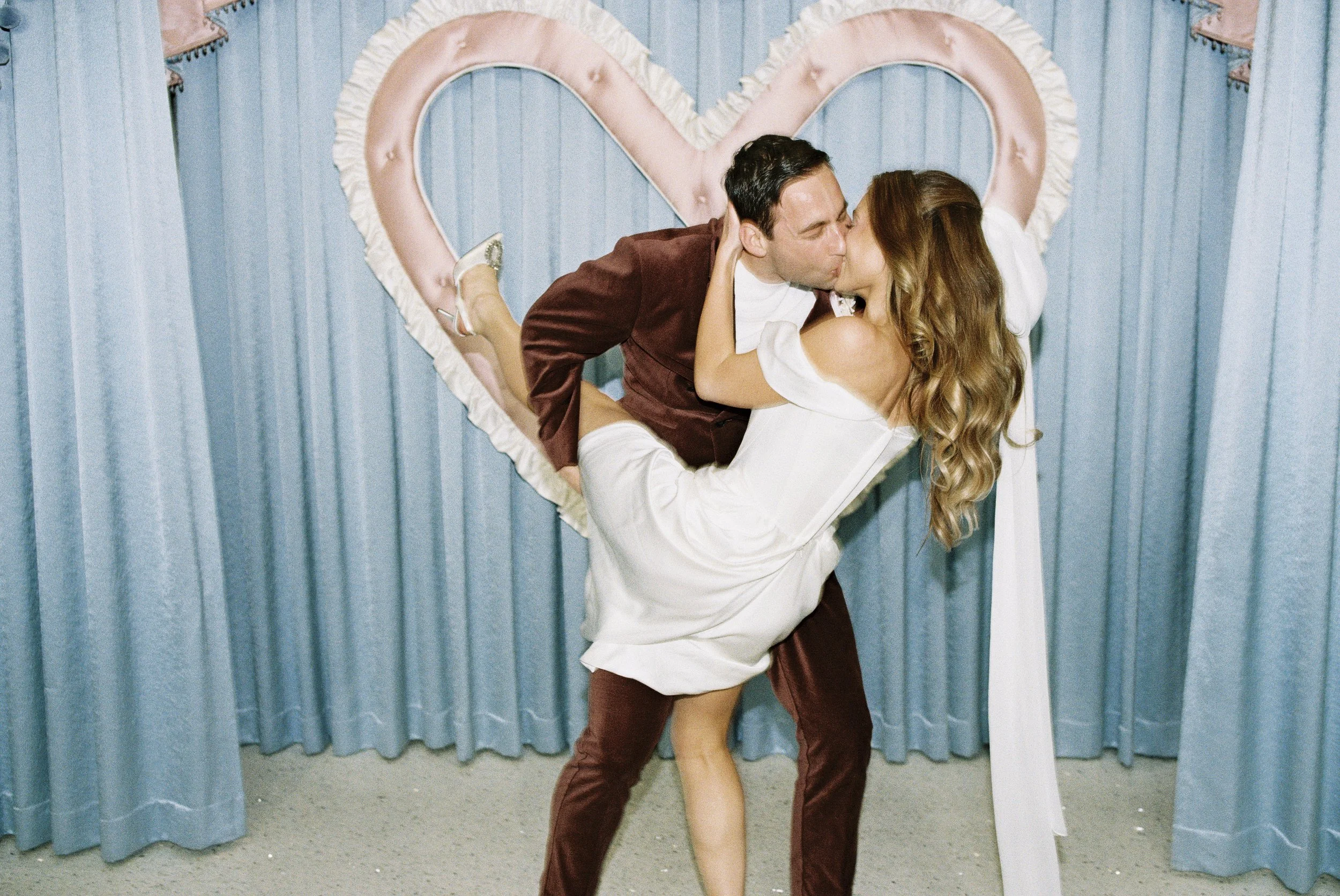 A couple sharing a passionate kiss, with the woman lifted off the ground by the man, in front of a blue curtain decorated with a pink heart-shaped outline.