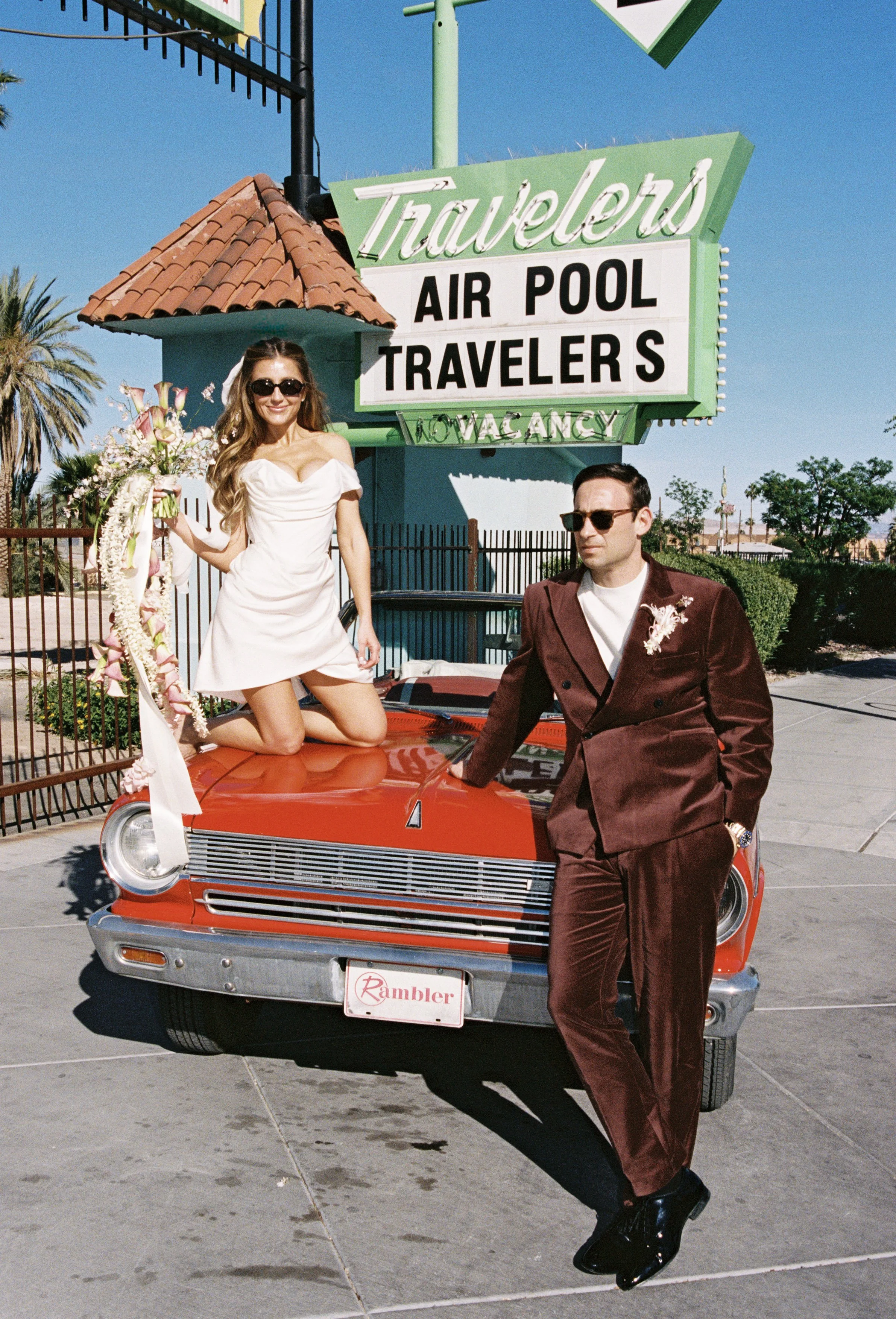 A man and woman pose outside next to a red vintage car in front of a sign that reads 'Travelers Air Pool Travelers' with a smaller sign underneath that says 'No Vacancy.' The woman is kneeling on the car with a bouquet of flowers, wearing a white dre