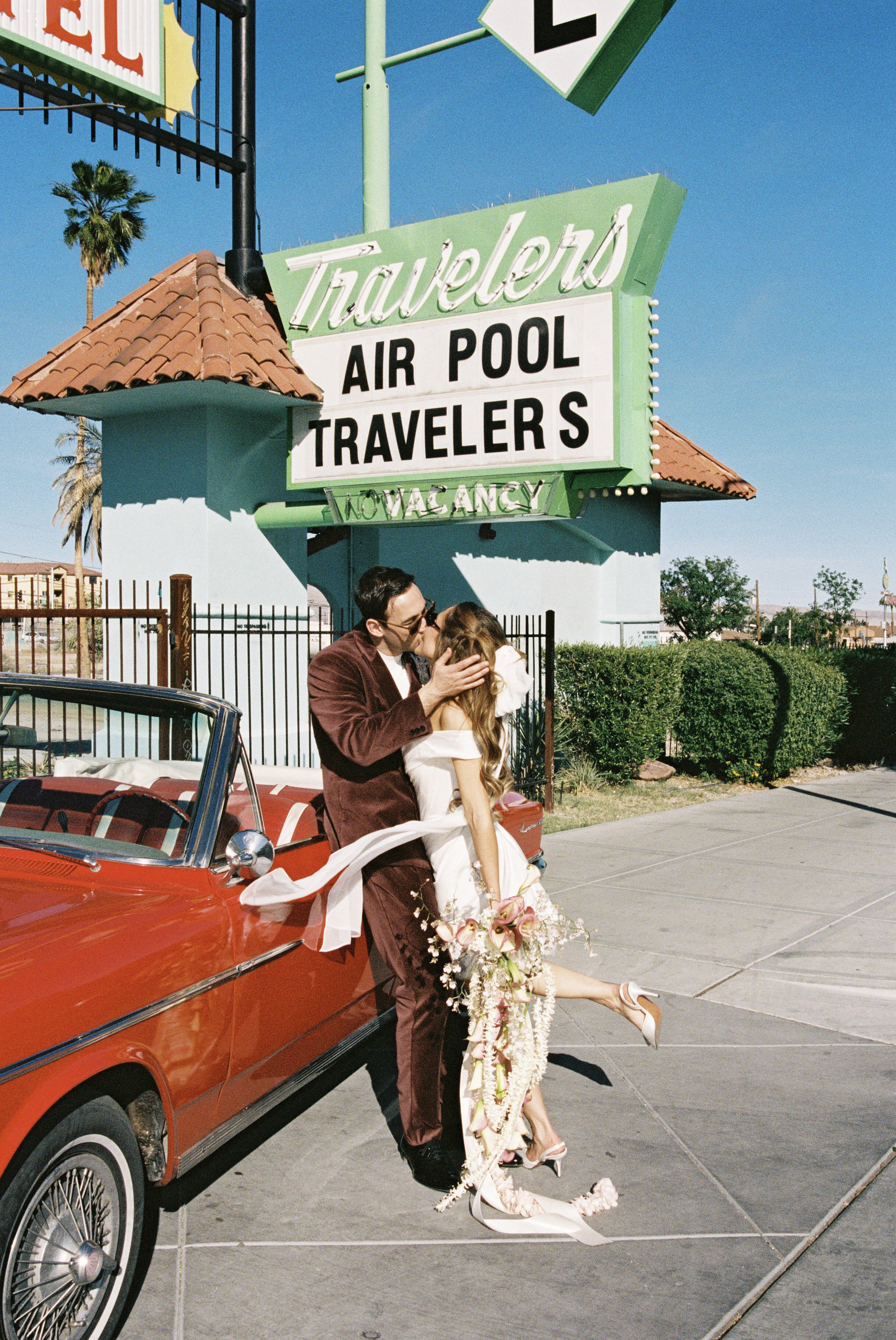 A couple in wedding attire kissing next to a red vintage car, with a large sign for 'Travelodge' in the background, under a clear blue sky.