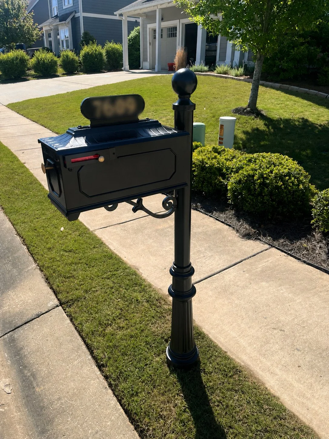 Black mailbox mounted on a decorative black post near the sidewalk in front of a house with a green lawn, shrubs, and trees.