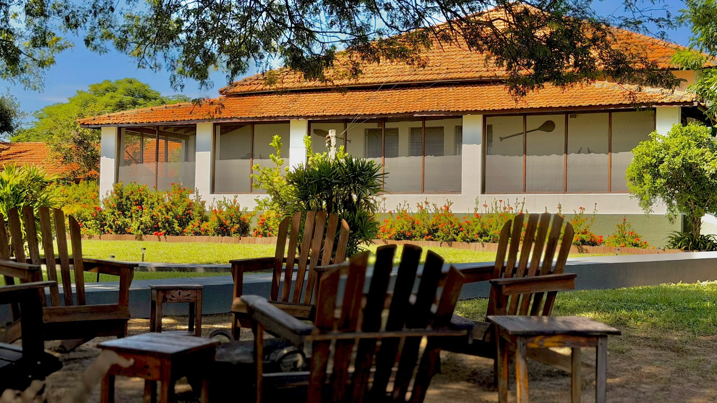 A view of a house with a red tile roof, white walls, and large windows, seen from a shaded outdoor patio with wooden chairs and a table, surrounded by green lawns and colorful flowering bushes.