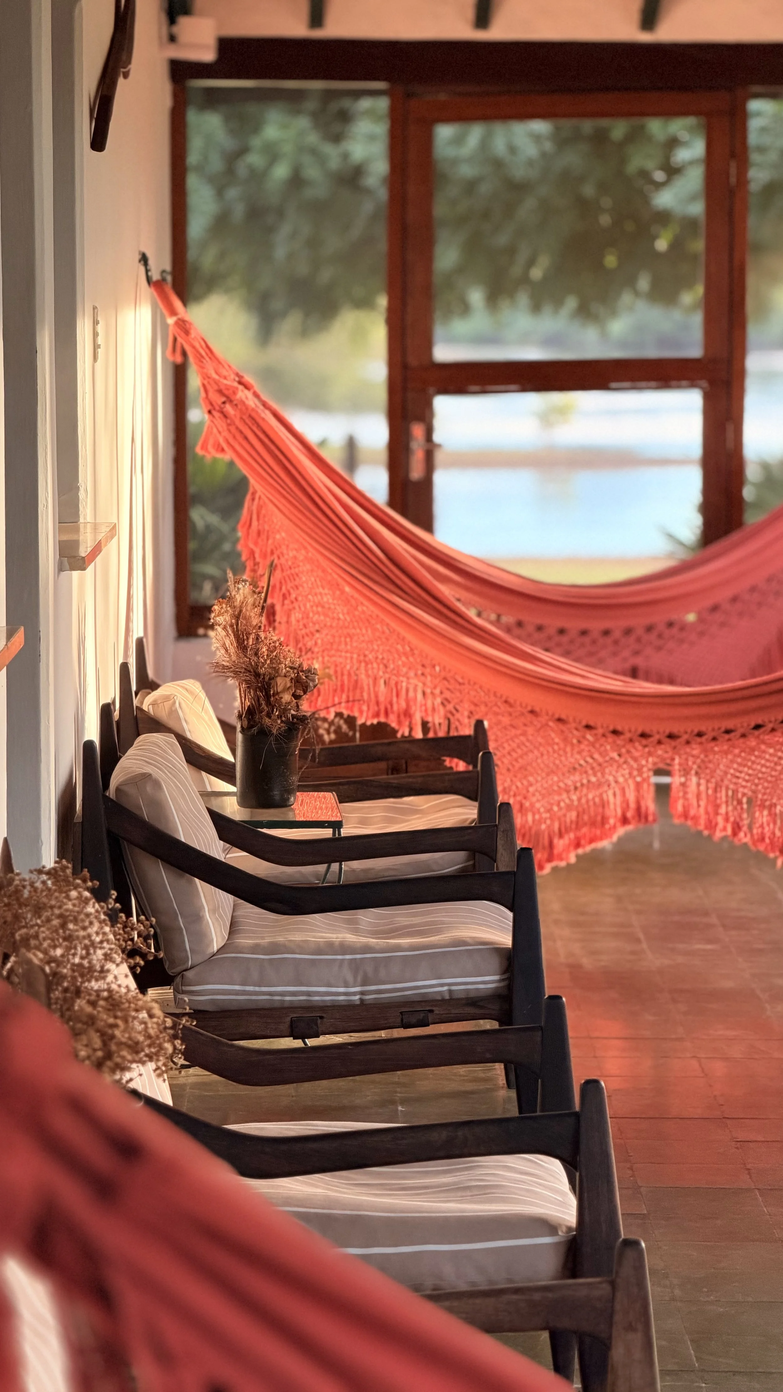 Interior of a cozy seating area with multiple wooden chairs with cushions, a black vase with dried flowers, and a large red hammock next to an open door or window with a view of greenery and water outside.