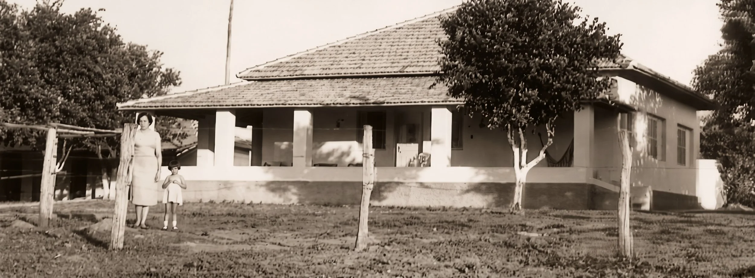 Black and white photo of a woman and a young girl standing in front of a house with a porch, trees, and a fence nearby.