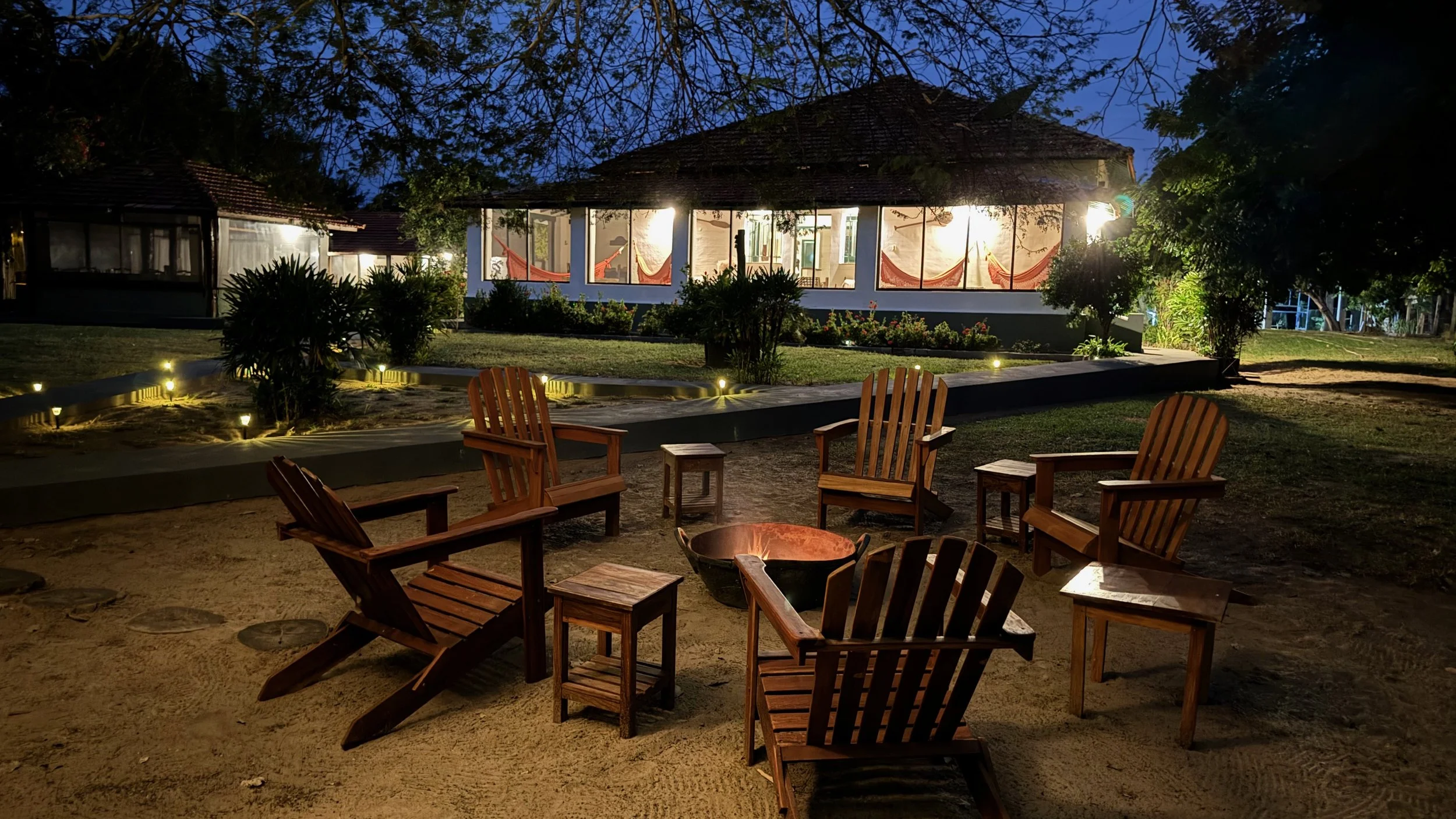 Nighttime view of a cozy outdoor seating area with wooden chairs around a fire pit, facing a glass-walled house illuminated from within, surrounded by trees and plants.