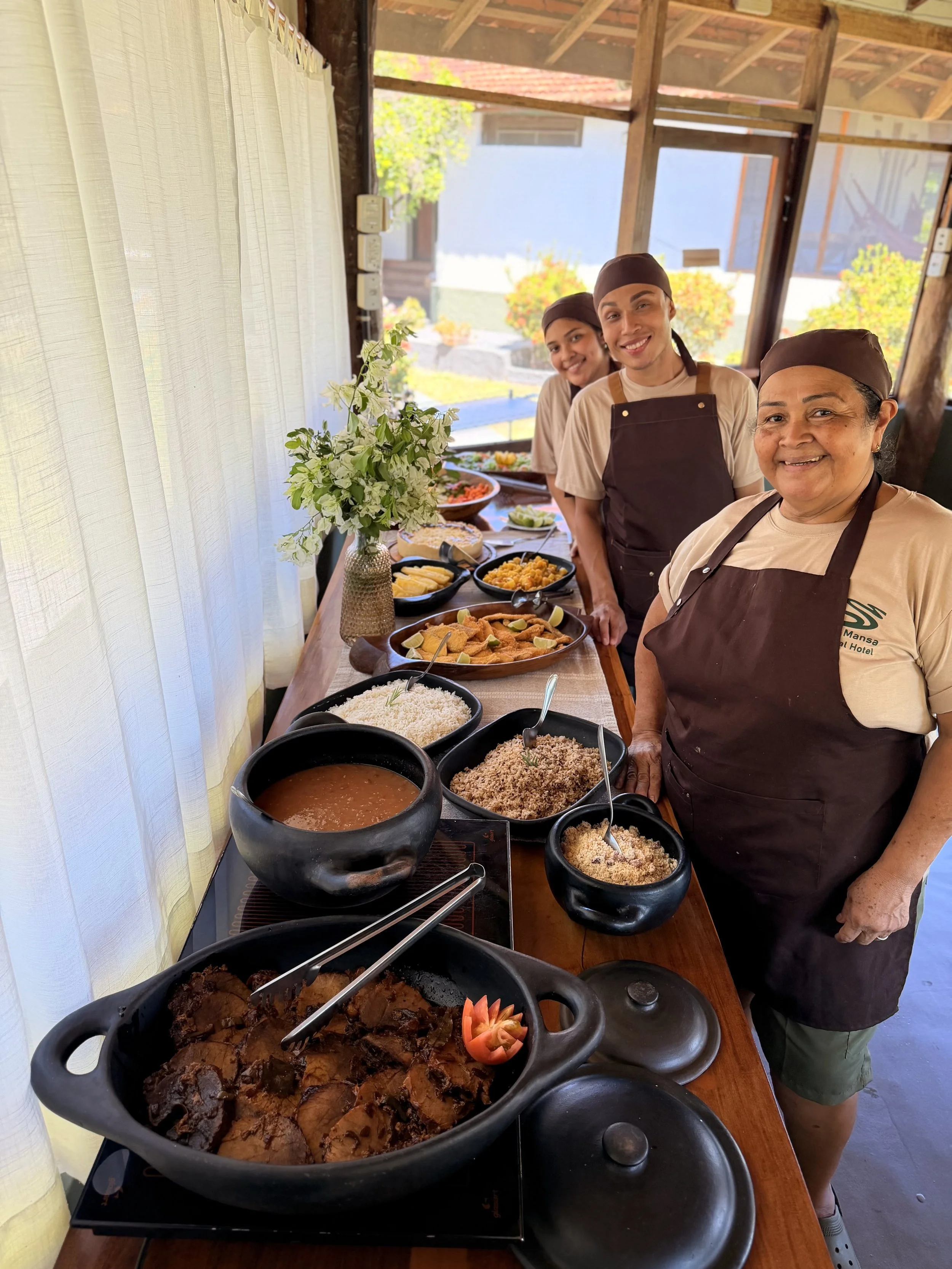 Three people in aprons and head coverings standing behind a table with various dishes, including rice, beans, and casseroles, in a room with large windows and white curtains.