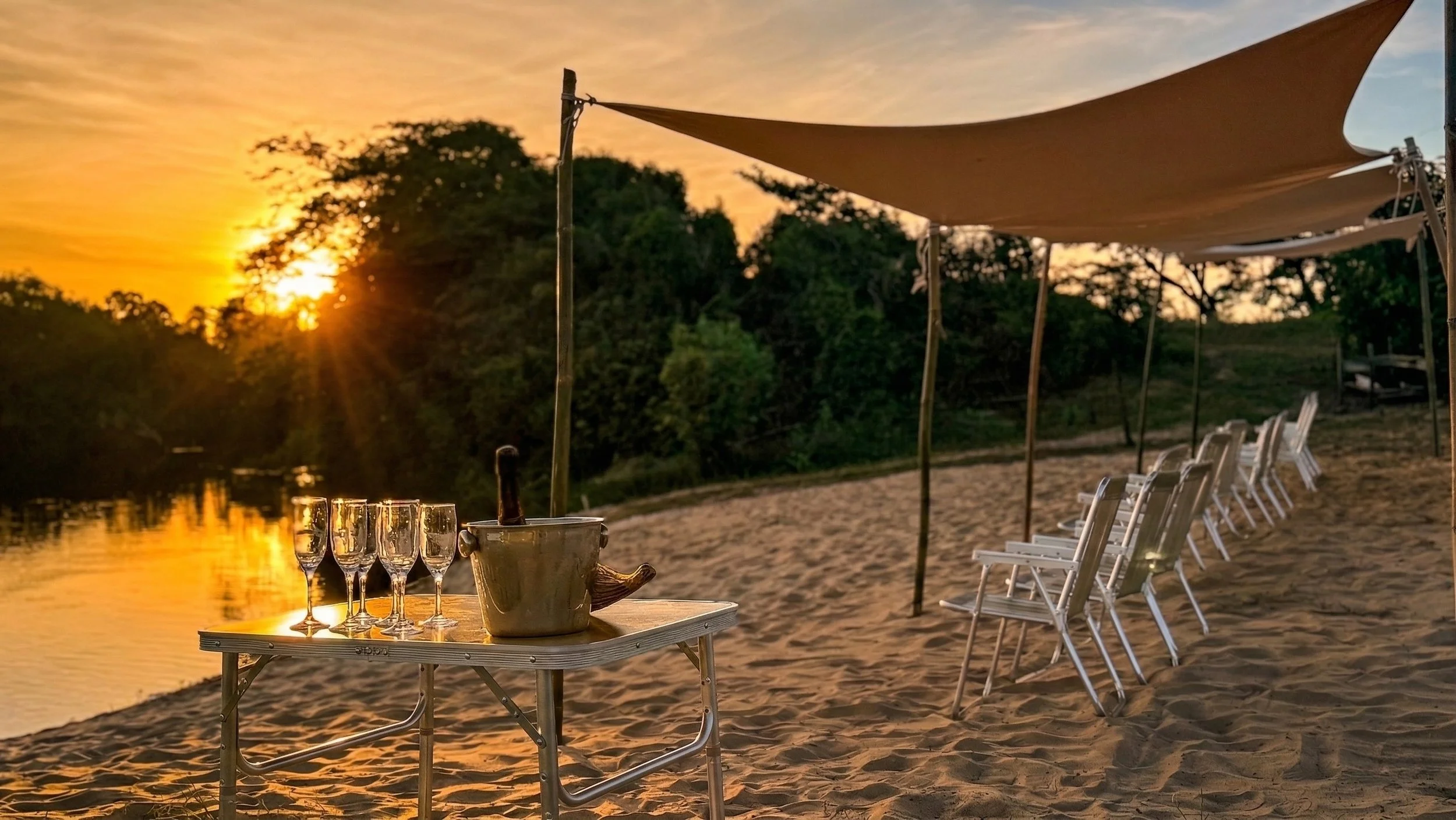 Outdoor setting at sunset with a table of glasses and a bucket, beach chairs under umbrellas along a sandy shore, calm water reflecting the sky, trees in the background.