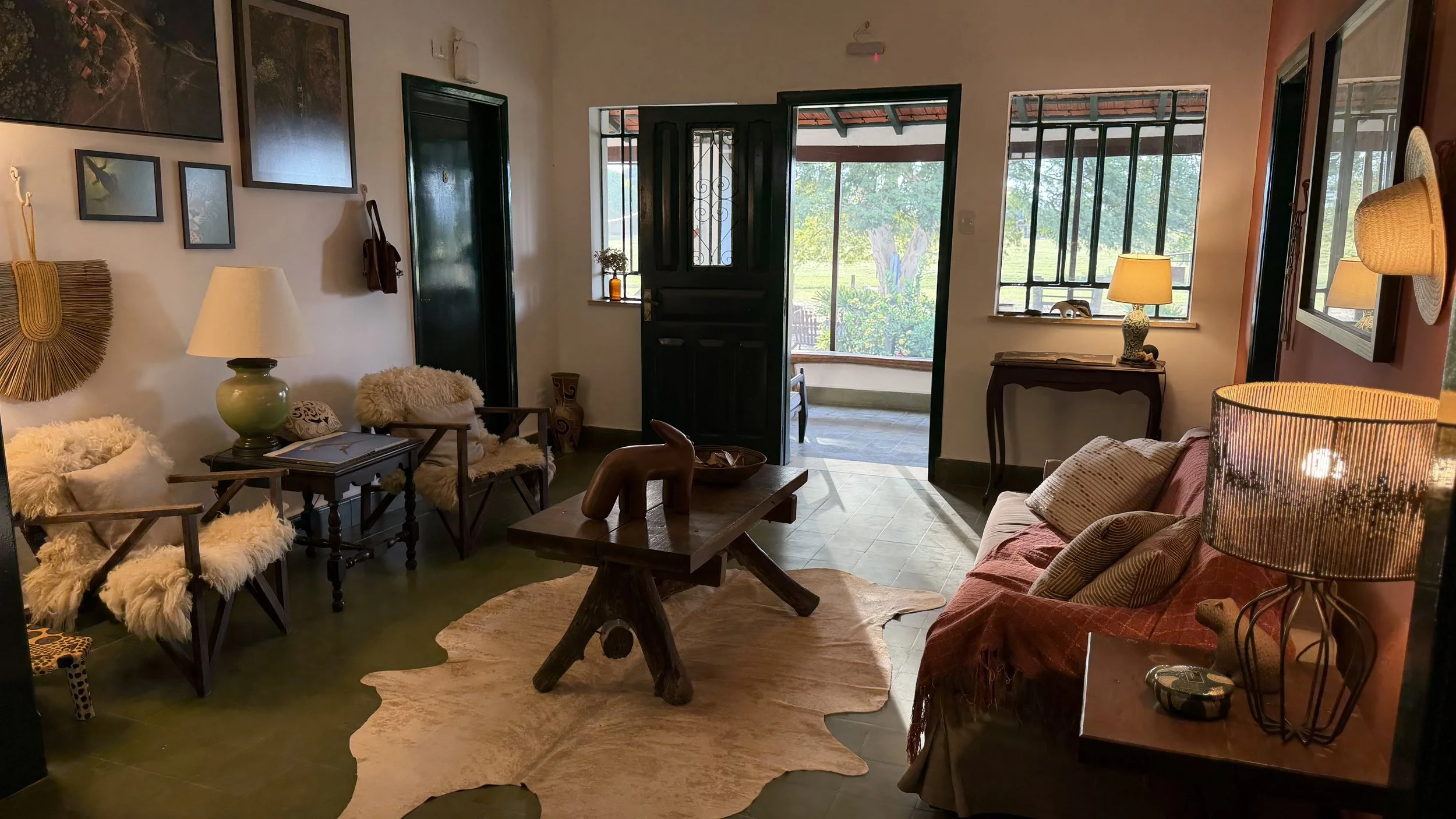 Cozy living room with two wooden armchairs with furry cushions, a sofa with striped pillows, a wooden coffee table, and lamps providing warm lighting. Artwork decorates the wall, and large windows and a door open to a green outdoor area.