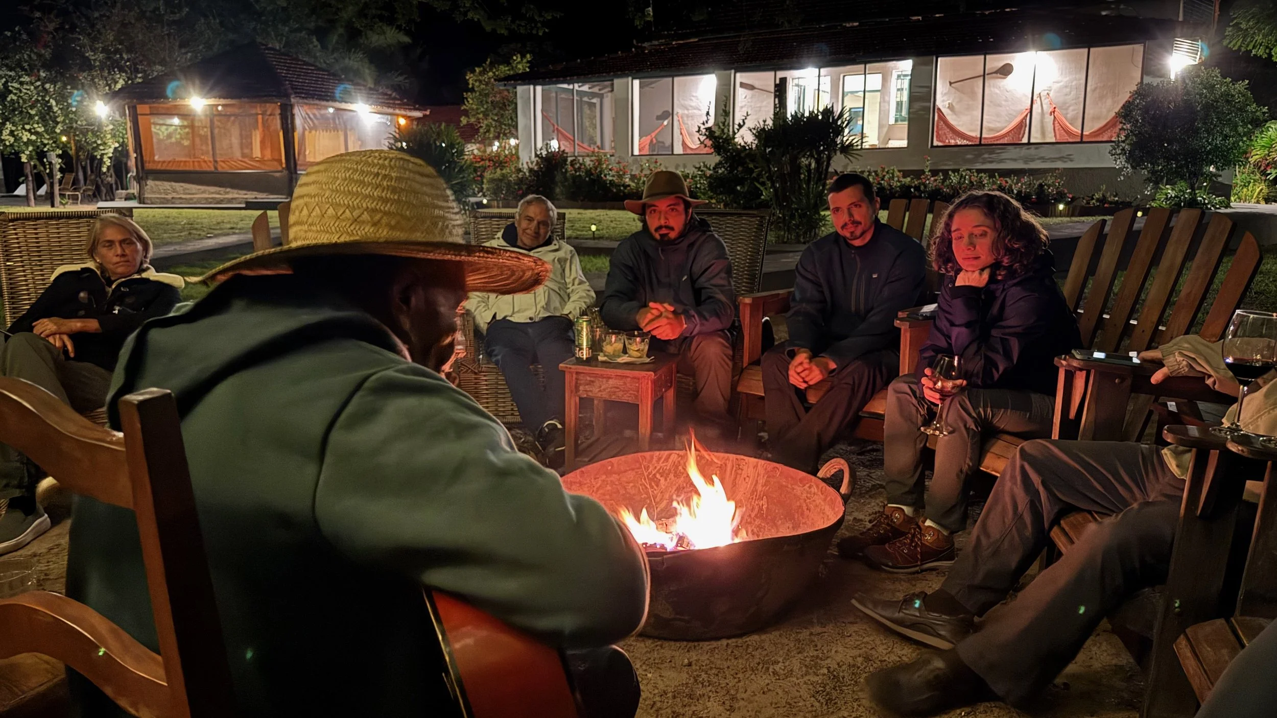 Group of people sitting outdoors at night around a fire pit, with a large windowed building and trees in the background