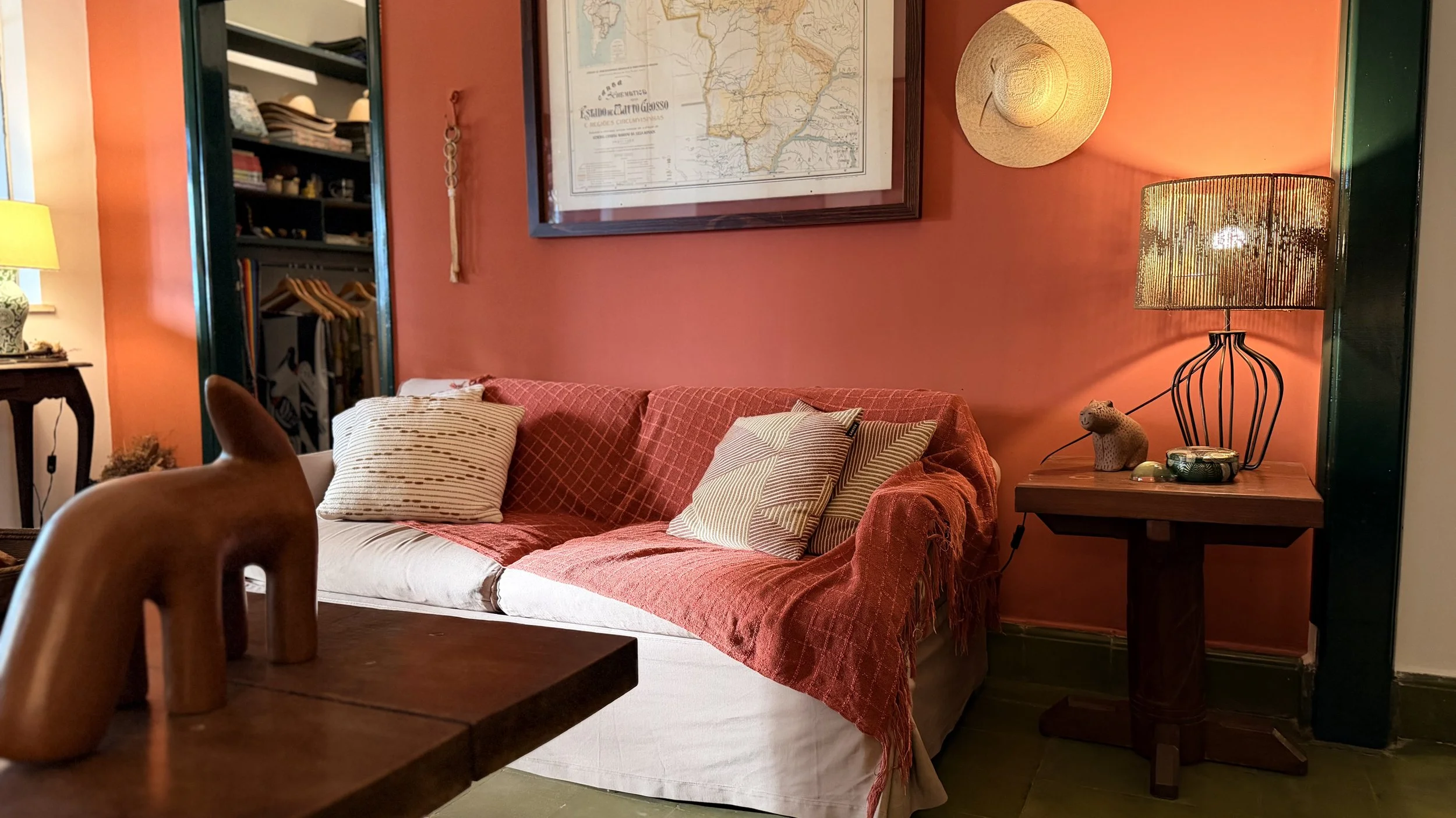 A cozy living room corner with a red sofa adorned with patterned throw pillows, a side table with a woven hat hung on the wall, a framed map, and a lamp with a woven shade providing warm lighting.