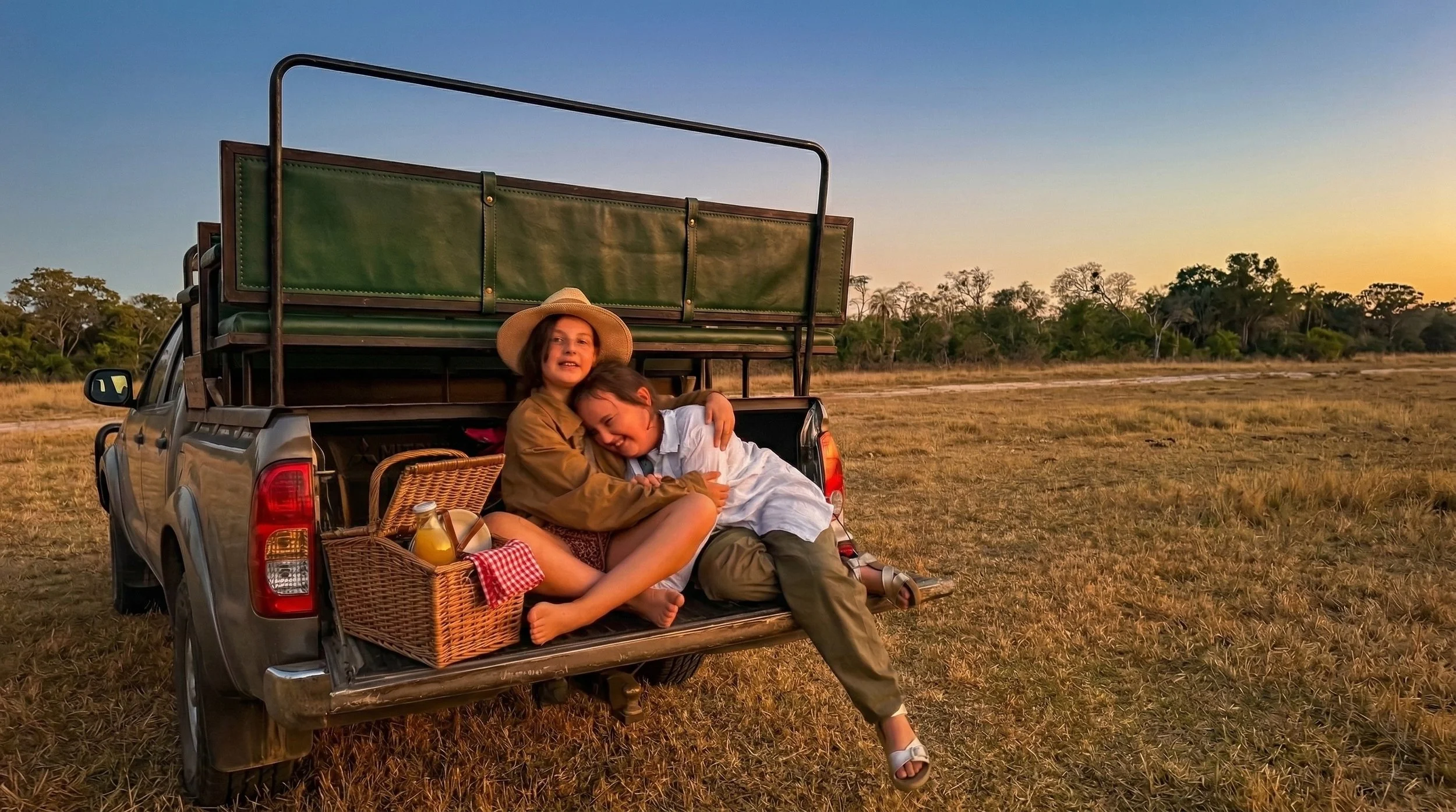 Two children sitting on the back of a pickup truck in a grassy field at sunset, hugging and smiling, with picnic supplies and a basket beside them.