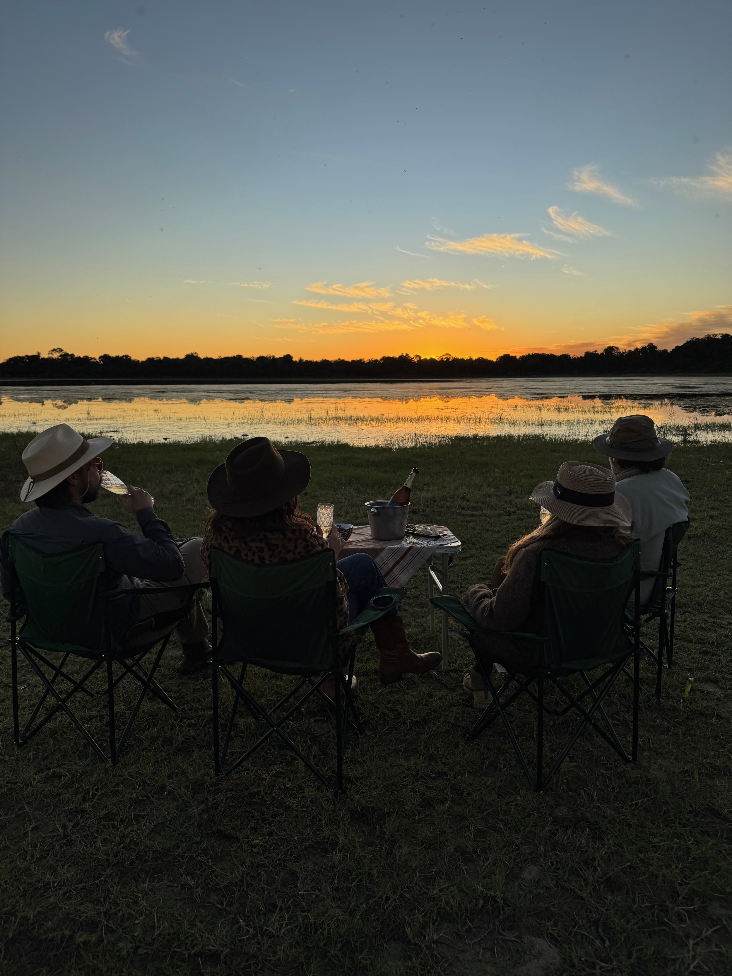People sitting on chairs outdoors watching a sunset over a river, with trees in the background, some wearing hats, a table with drinks in front of them.