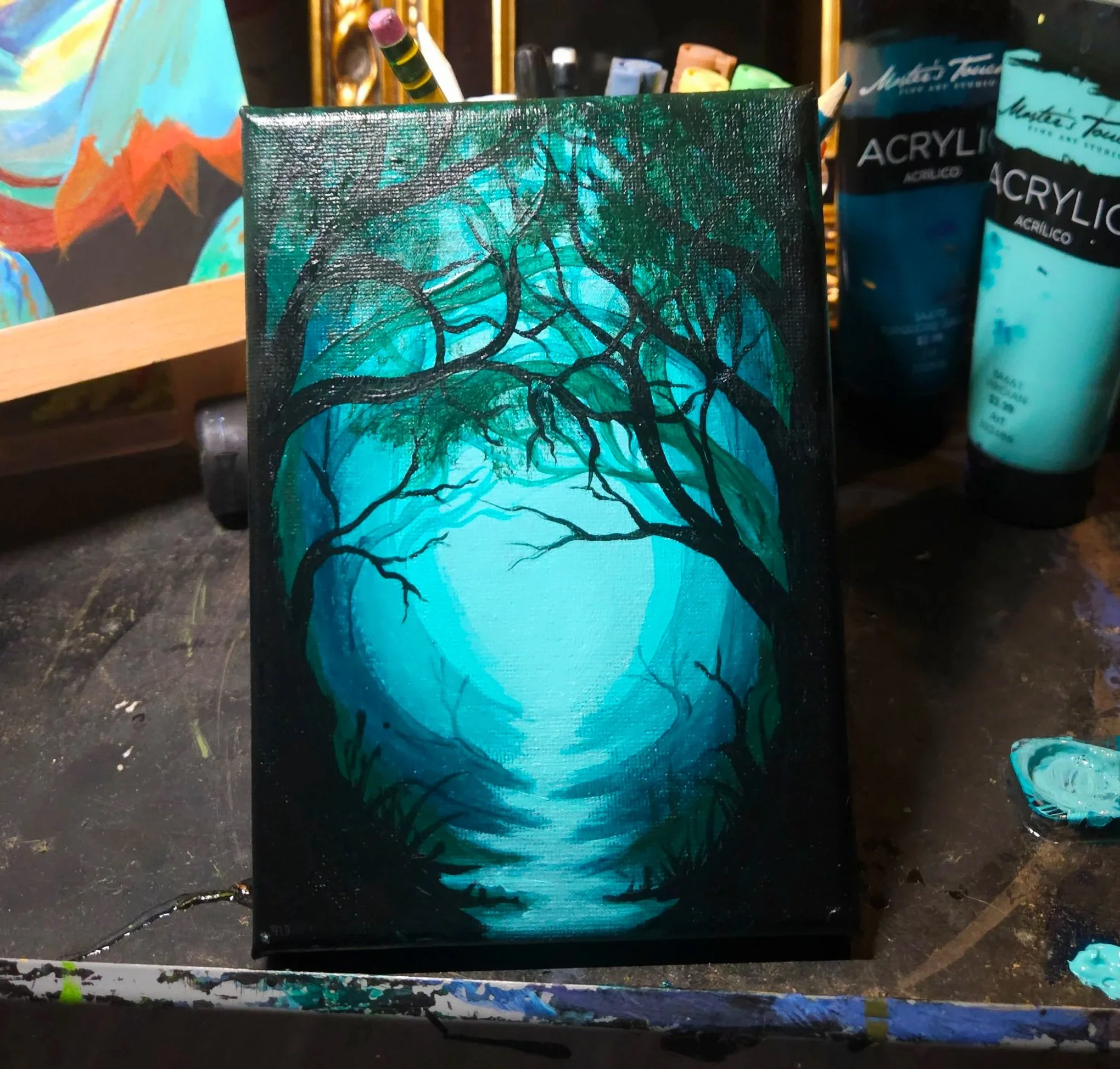 A small canvas with an abstract painting of a tree with black branches and teal and blue background, standing on a cluttered artist's workspace with various art supplies around.