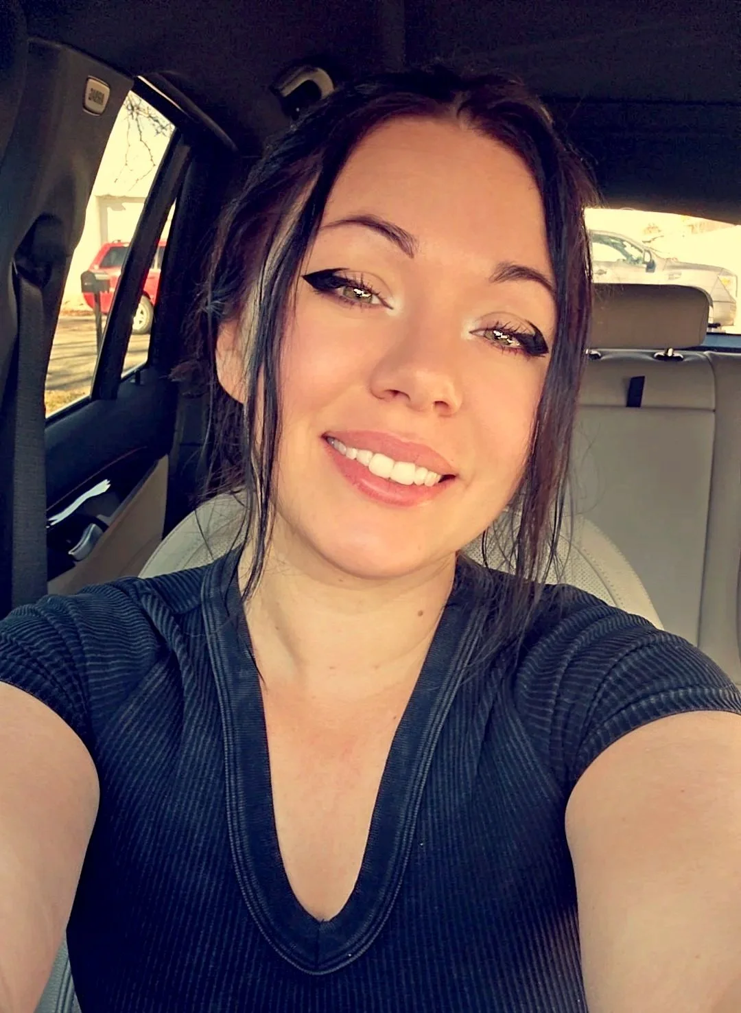 Young woman with dark hair in a ponytail, wearing eyeliner and a dark shirt, taking a selfie inside a car with a light-colored interior, parked outdoors with other cars visible through the windows.