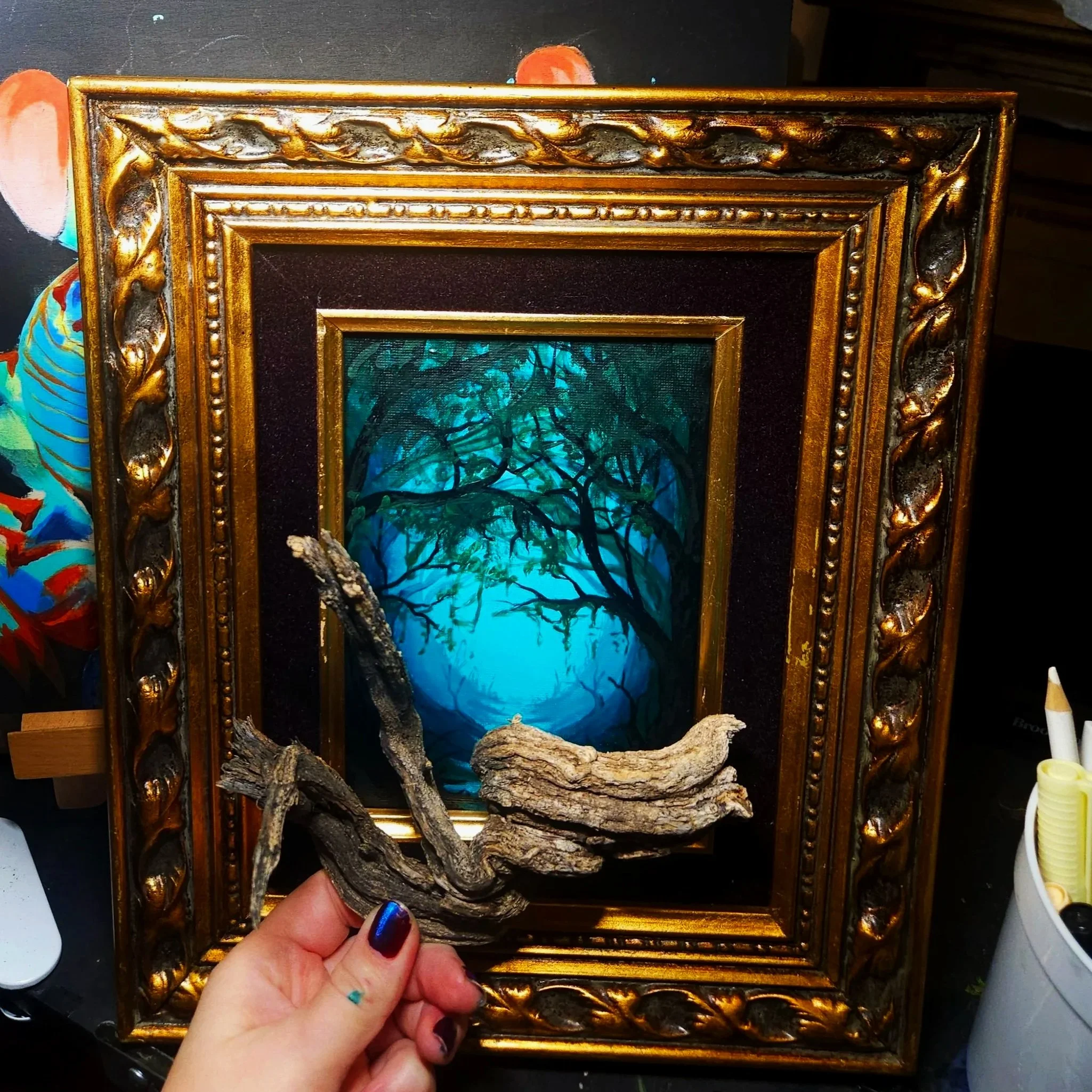Close-up of a decorative, ornate gold picture frame being held by a person's hand. The frame has intricate carvings and a shiny finish. The background inside the frame features a vibrant, blue-colored image of a tree with branches, giving a painting-like appearance.