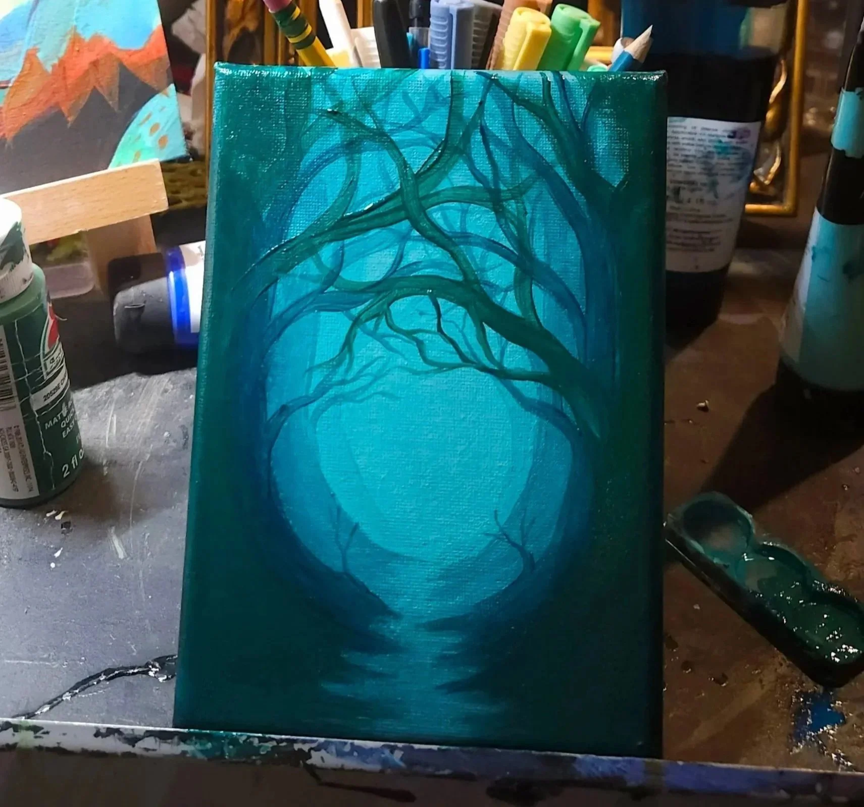 A painting in progress featuring a blue background with dark blue and green tree branches. Painting supplies, including brushes and paints, are visible around the artwork.