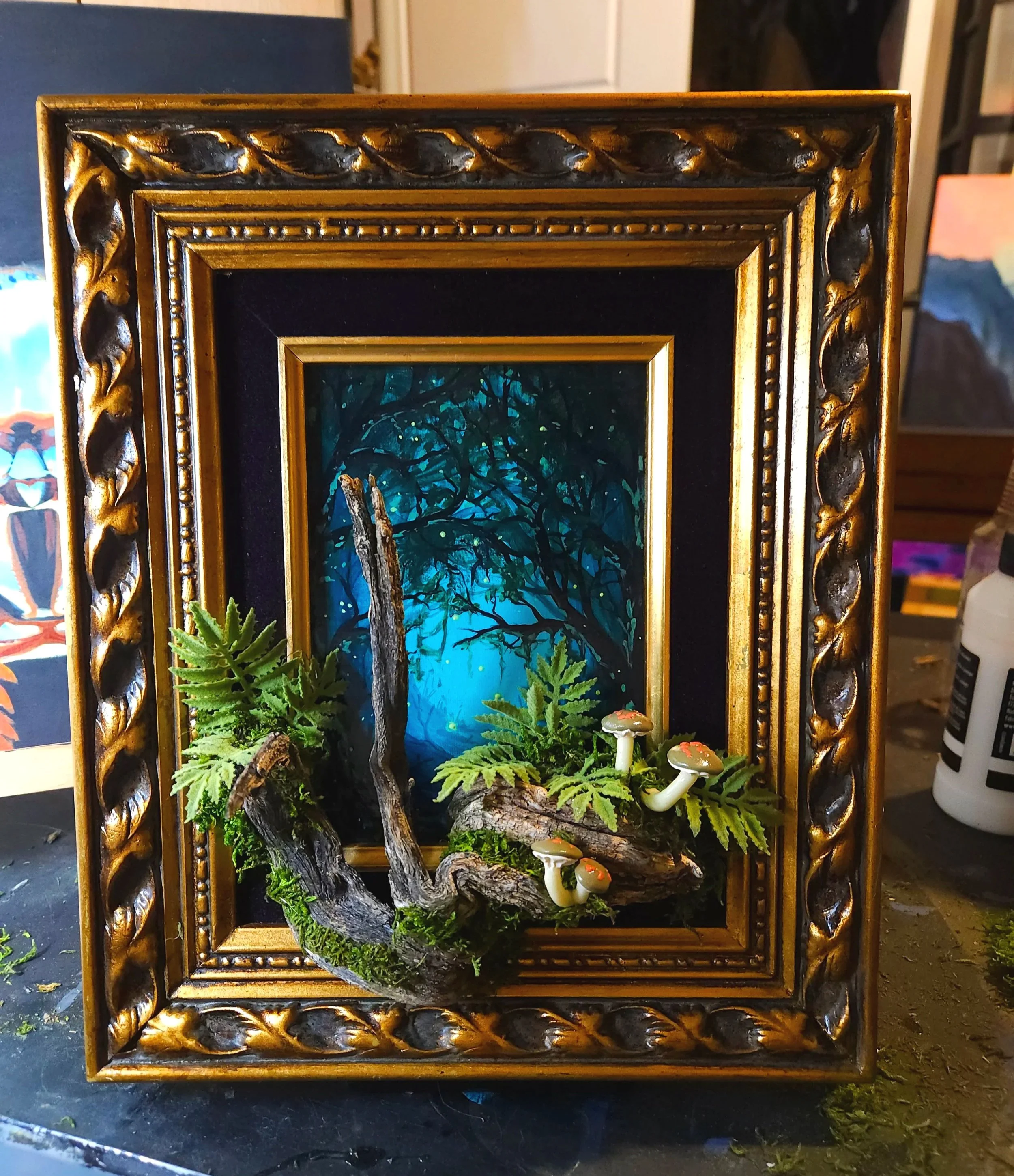 A decorative framed miniature landscape scene featuring a blue sky, a leafless tree, green plants, and small fungi.
