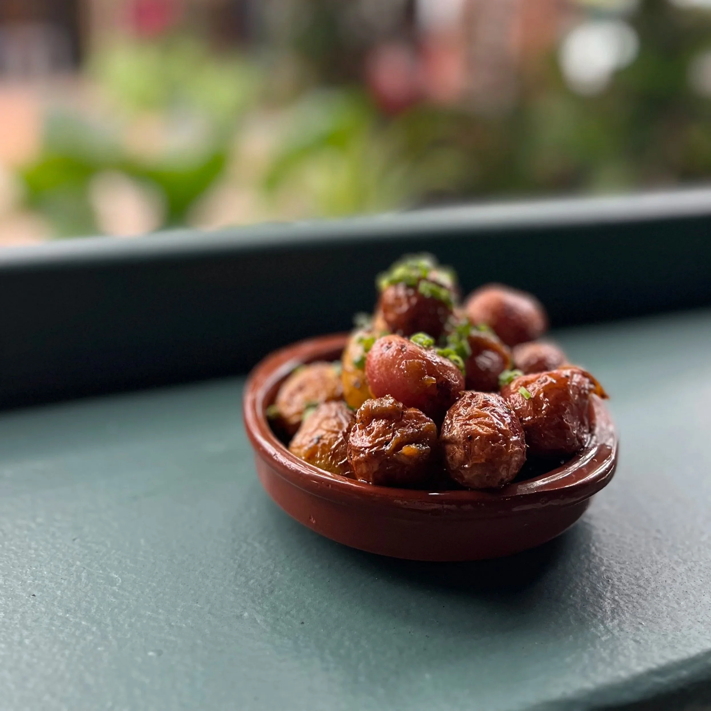 A small terracotta bowl filled with cooked chickpeas garnished with chopped green herbs, placed on a dark green surface with a blurred outdoor background.