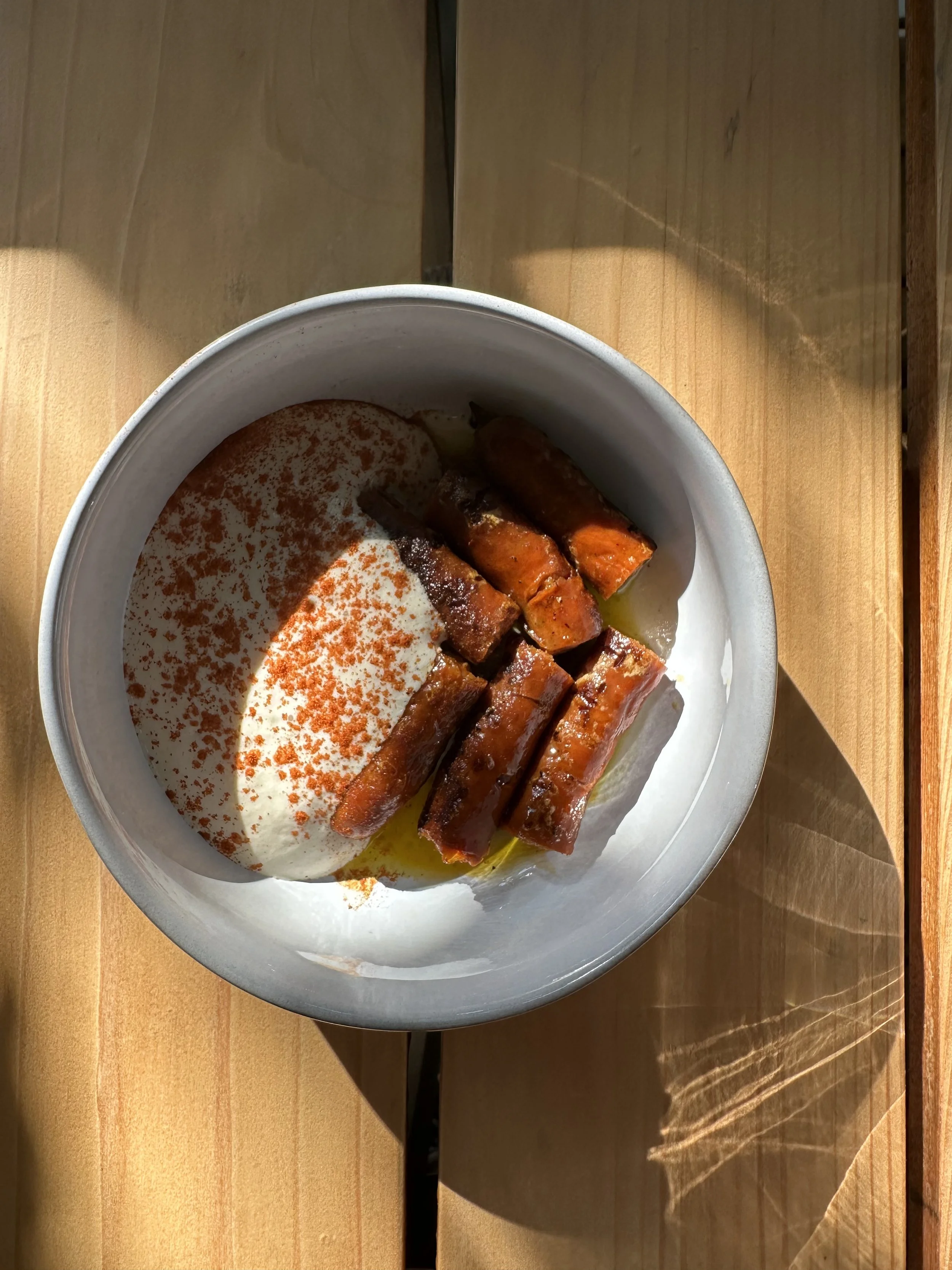 Bowl of yogurt topped with red pepper flakes, served with sliced roasted sweet potatoes or carrots, on a wooden table.