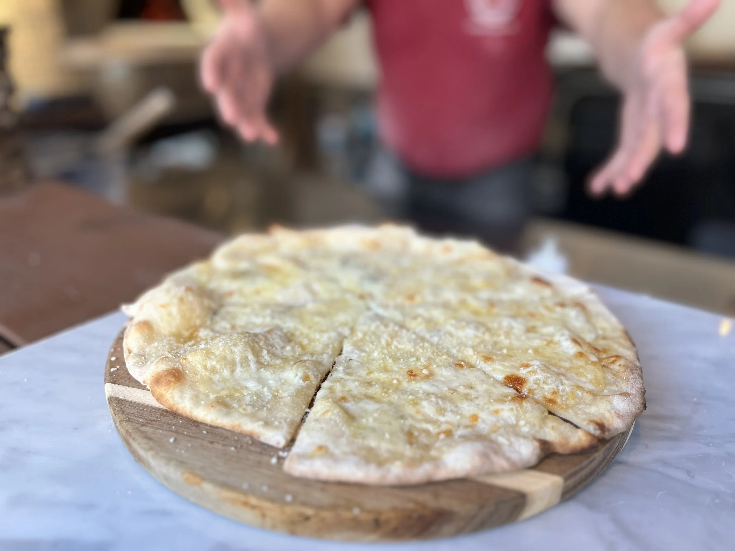 Cheese pizza on a wooden serving board with a blurred person in the background.