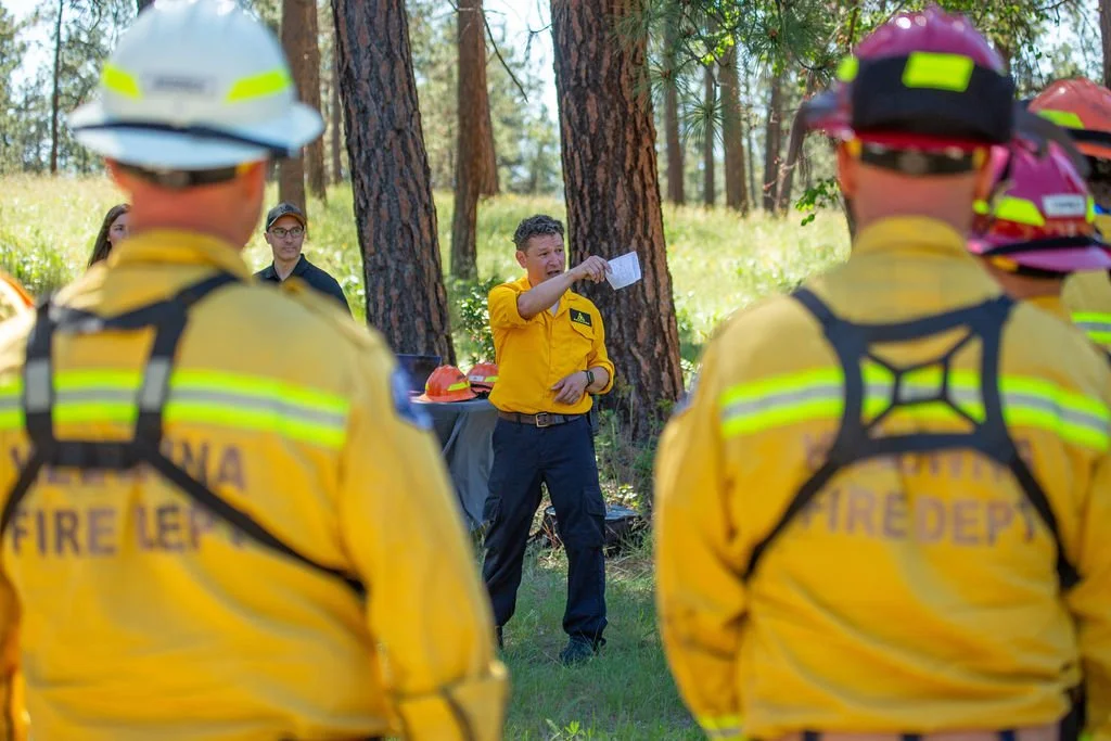 Firefighters in yellow uniforms listening to a FireSwarm team giving a safety briefing in a wooded outdoor area.