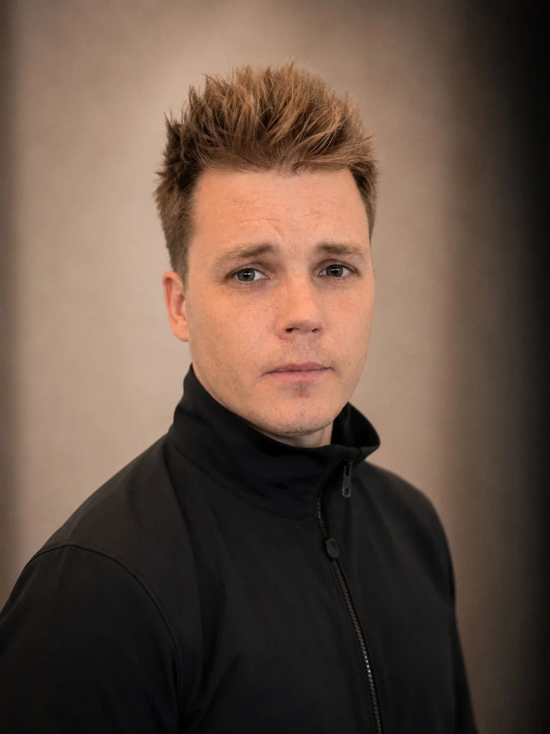 A man with short, styled brown hair and light skin wearing a black zip-up jacket looks at the camera against a neutral background.
