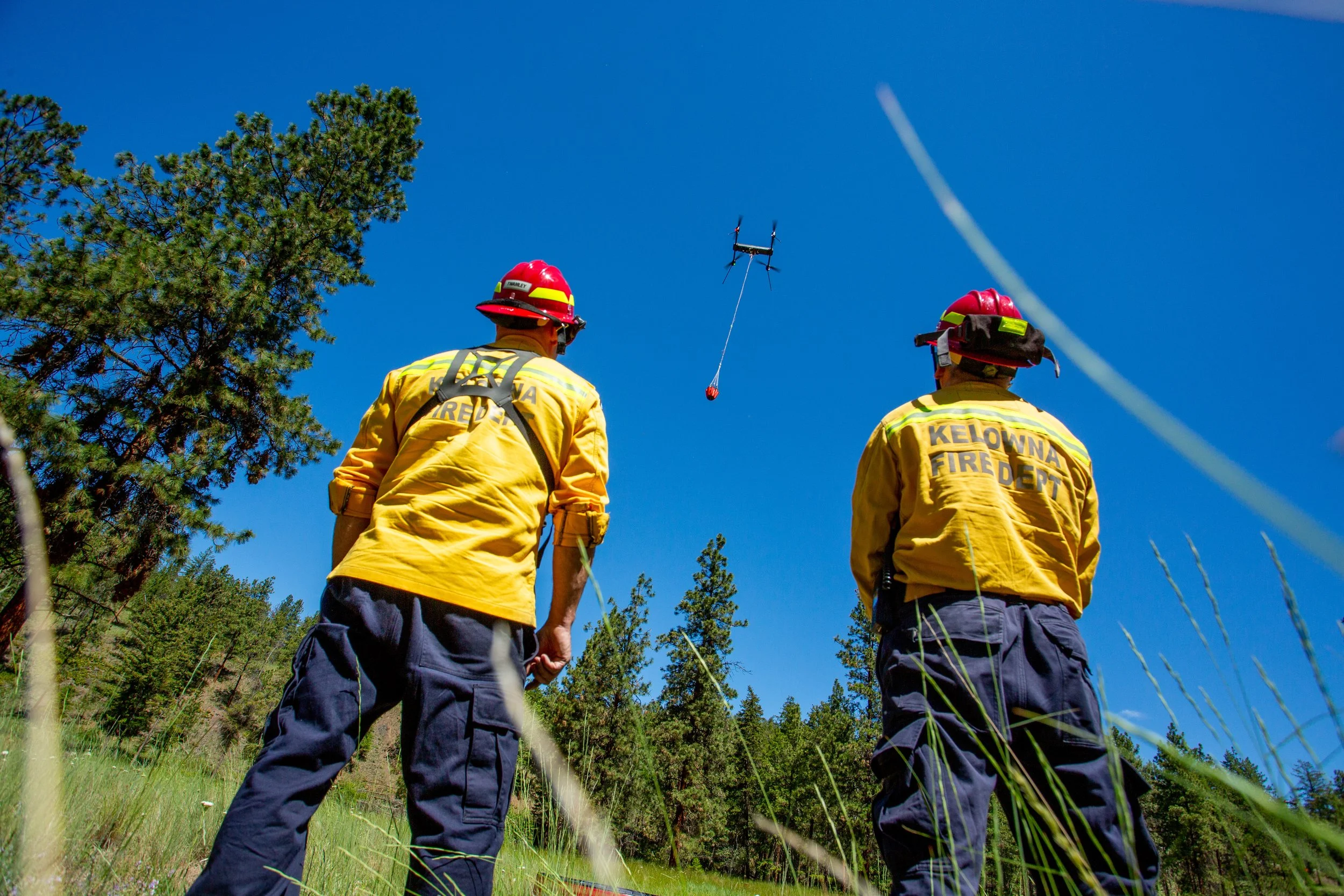 Two firefighters in yellow uniforms and red helmets observe the sky while a drone is flying overhead, attached to a cable or tether, in a wooded area under a clear blue sky.