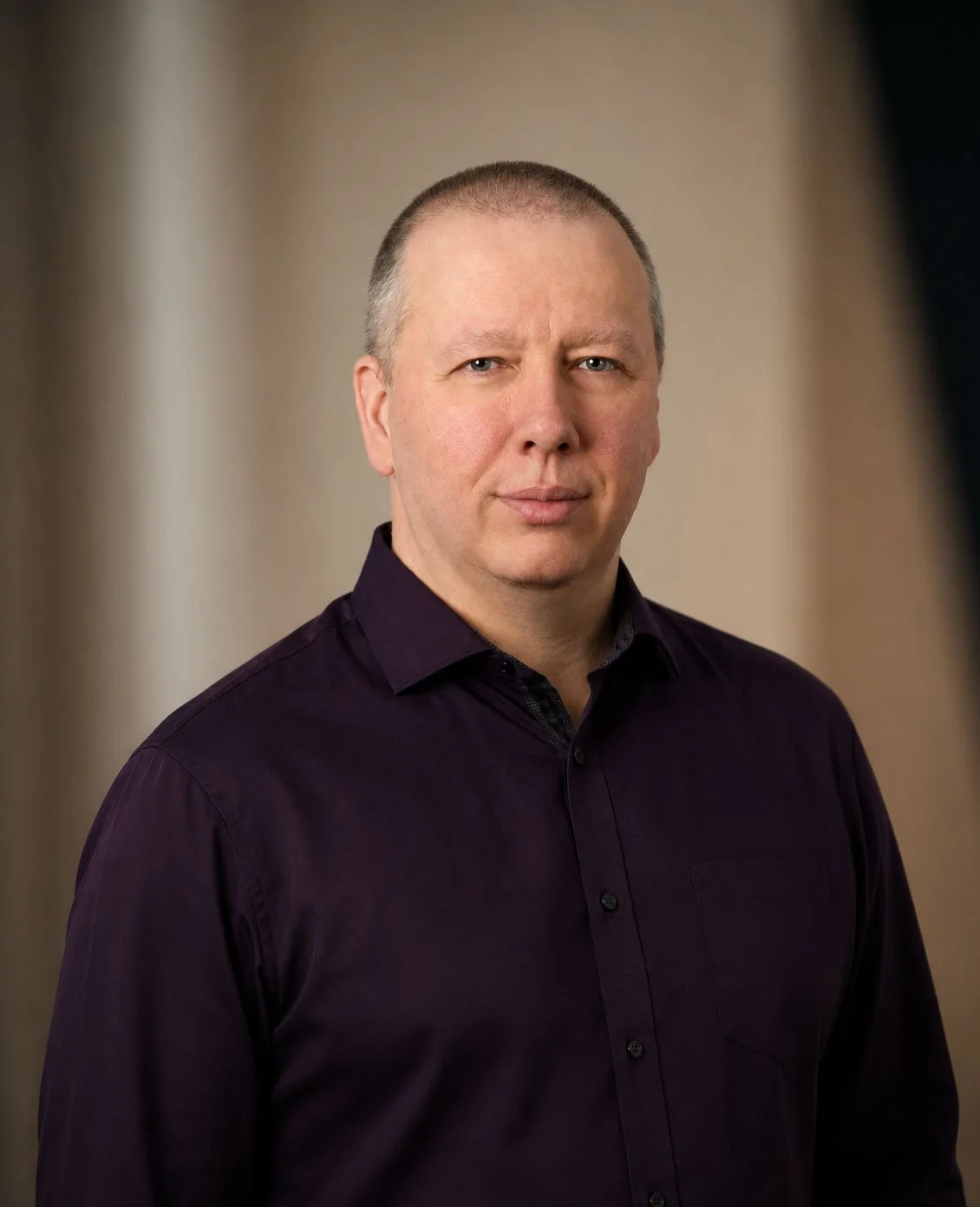 A man with short hair and a serious expression, wearing a dark purple button-up shirt, standing against a blurred neutral background.