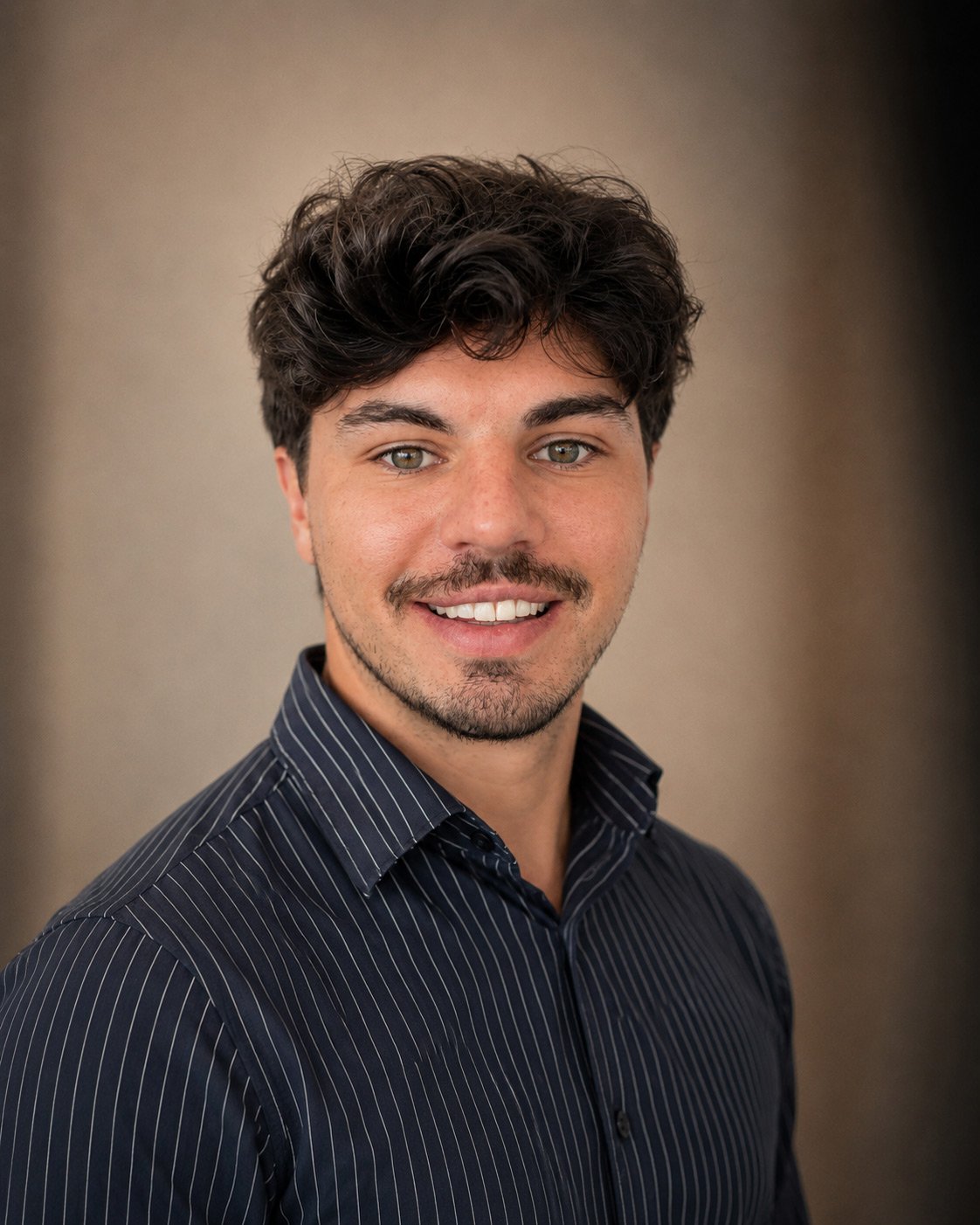 A young man with dark, wavy hair and light facial hair, wearing a dark navy, pinstriped dress shirt, smiling against a neutral background.