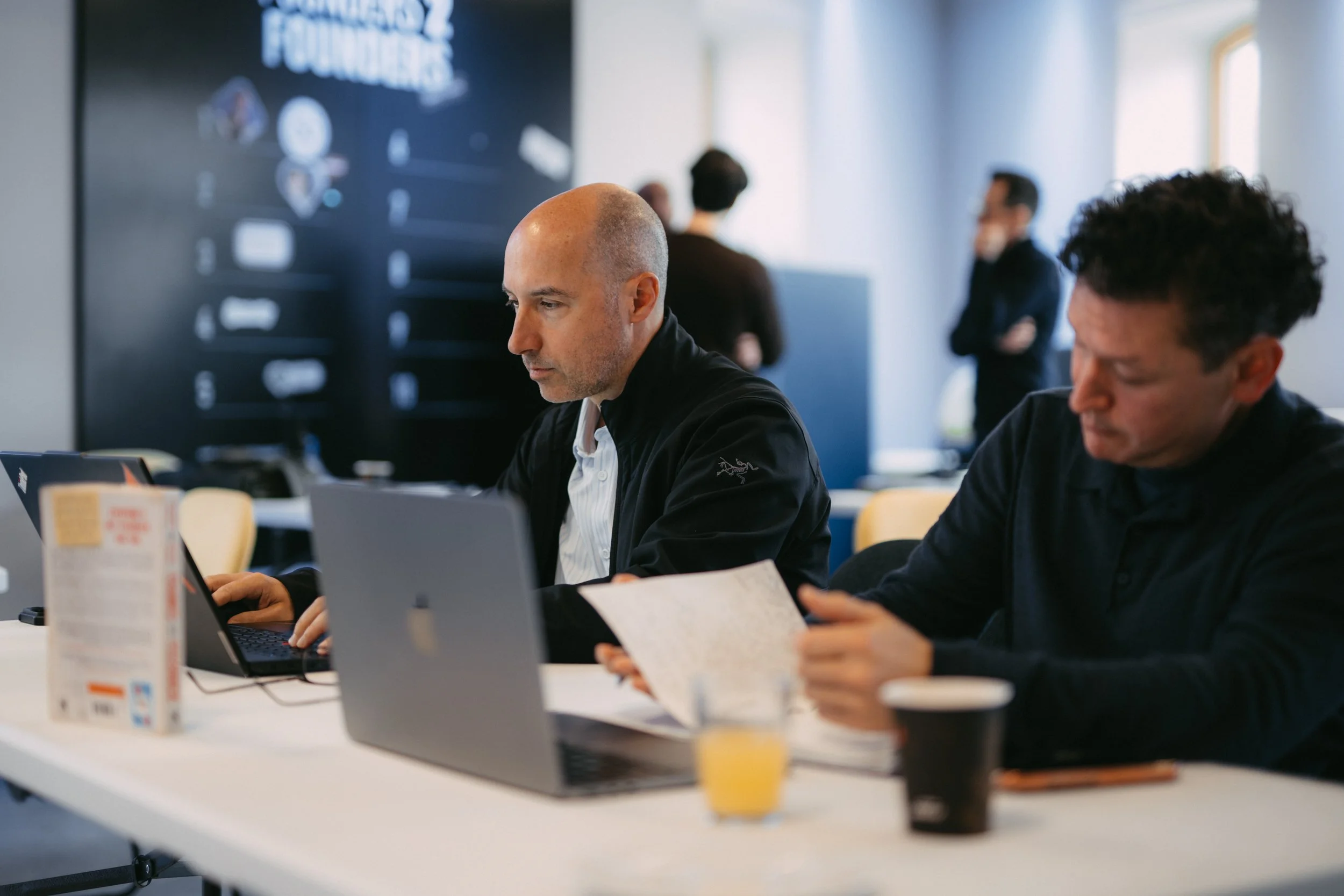 FireSwarm team working on laptops at a white table in a bright room with window light, with cups and snacks in front of them, and other people in the background.