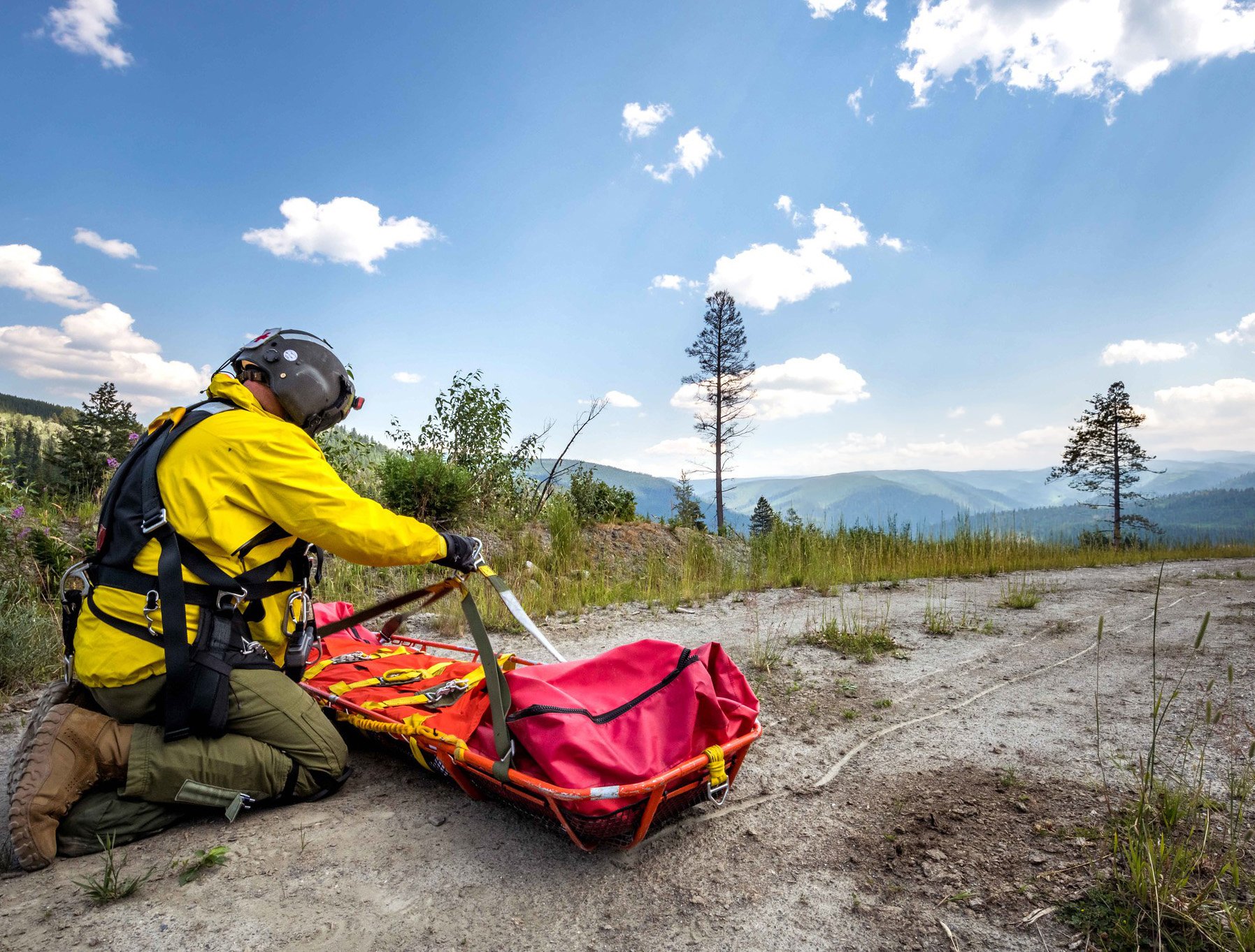 Person in yellow jacket with helmet kneeling next to a red rescue stretcher in a mountainous outdoor setting with trees and a blue sky.