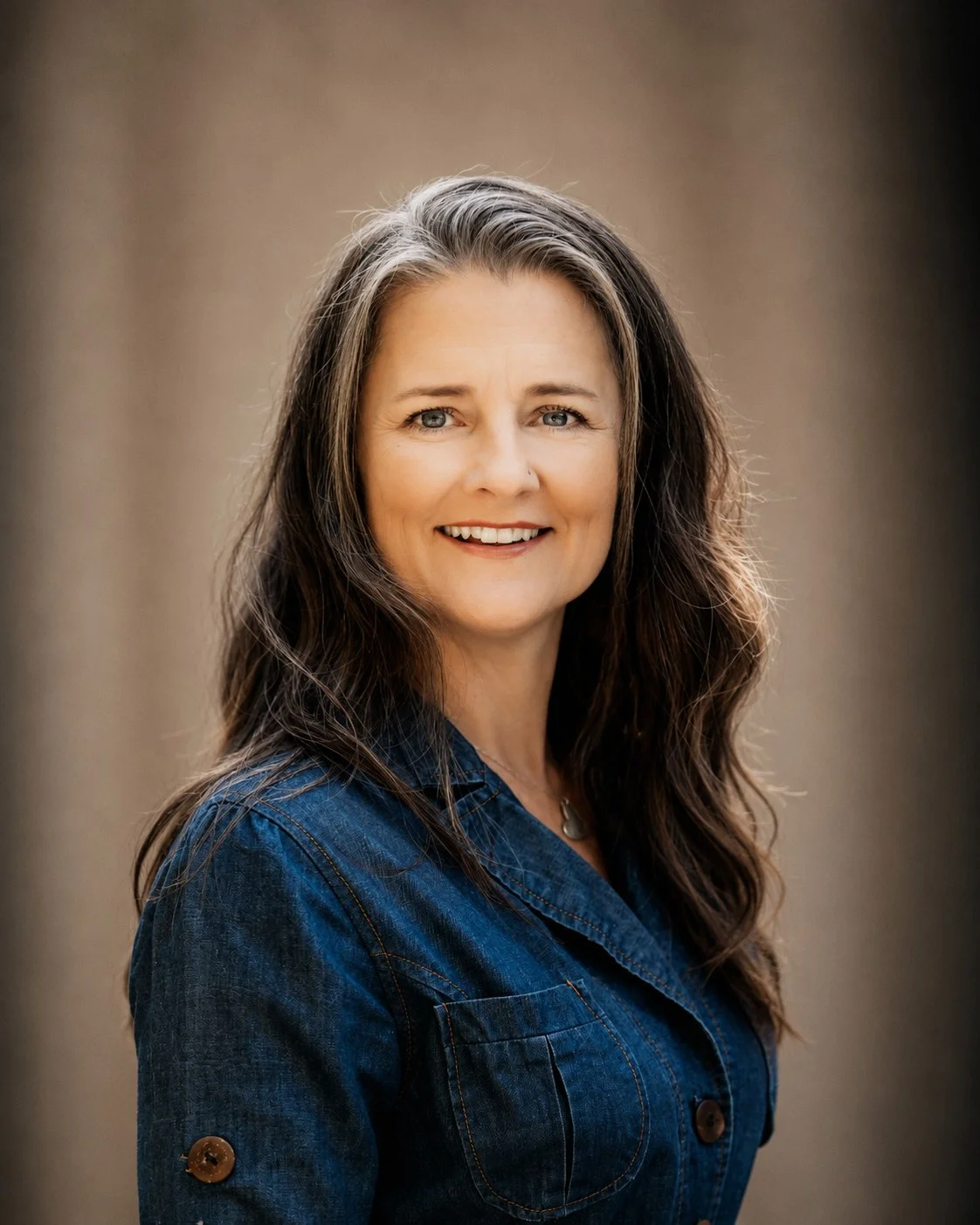 A woman with long brown hair and blue eyes smiling, wearing a denim jacket, outdoors with a blurred neutral background.