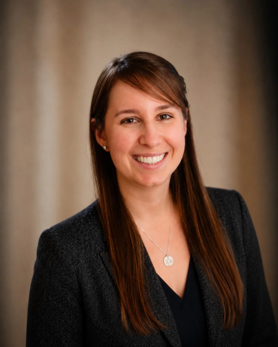 A smiling woman with brown hair and light skin, wearing a black blazer, a necklace, and earrings, posed against a neutral background.