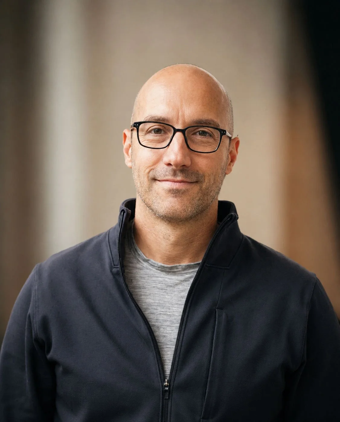 A smiling middle-aged man with glasses, a beard, and a shaved head, wearing a dark jacket and a gray shirt, standing indoors.