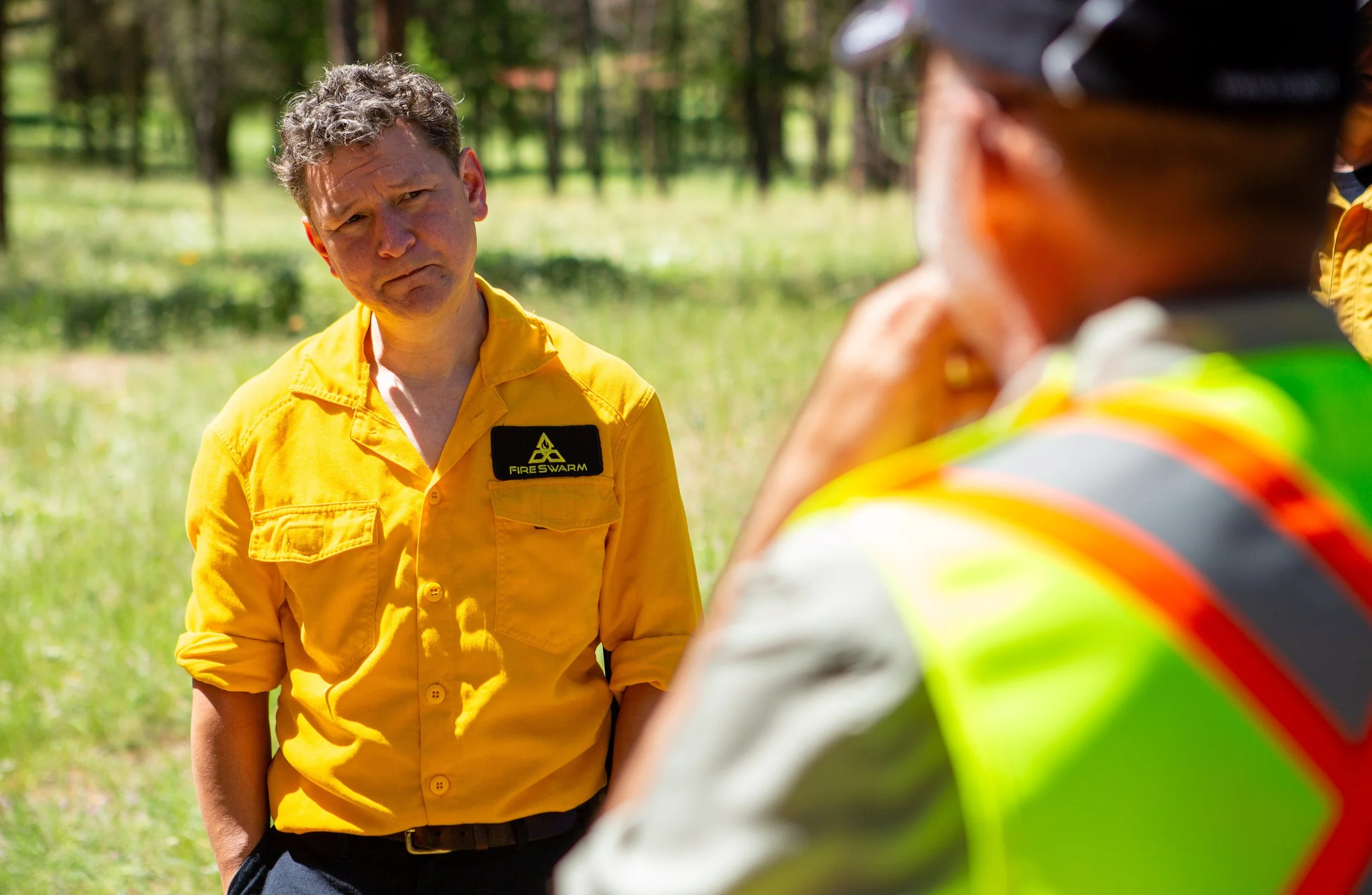 A man in a yellow uniform with a 'Fire Swarm' patch looks serious while talking to another person in a reflective vest and helmet outdoors on a sunny day, with a background of trees and grass.