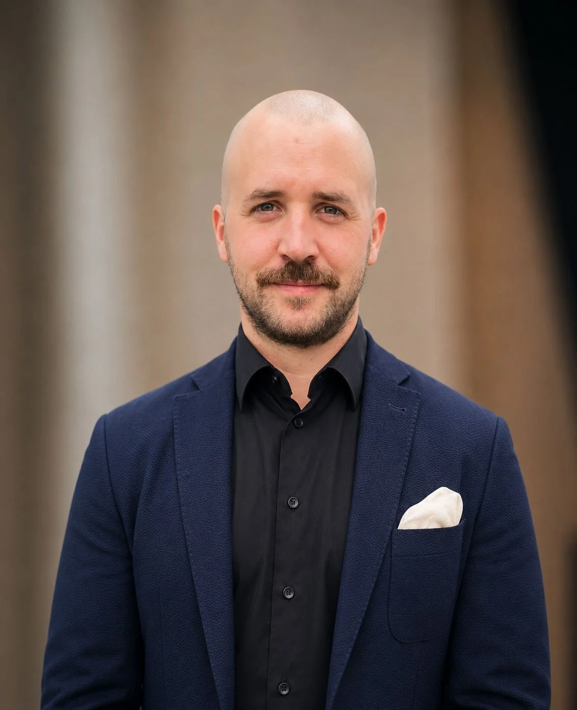 Headshot of a man with a bald head and beard, wearing a navy blazer, black shirt, and white pocket square, standing in front of a blurred background.