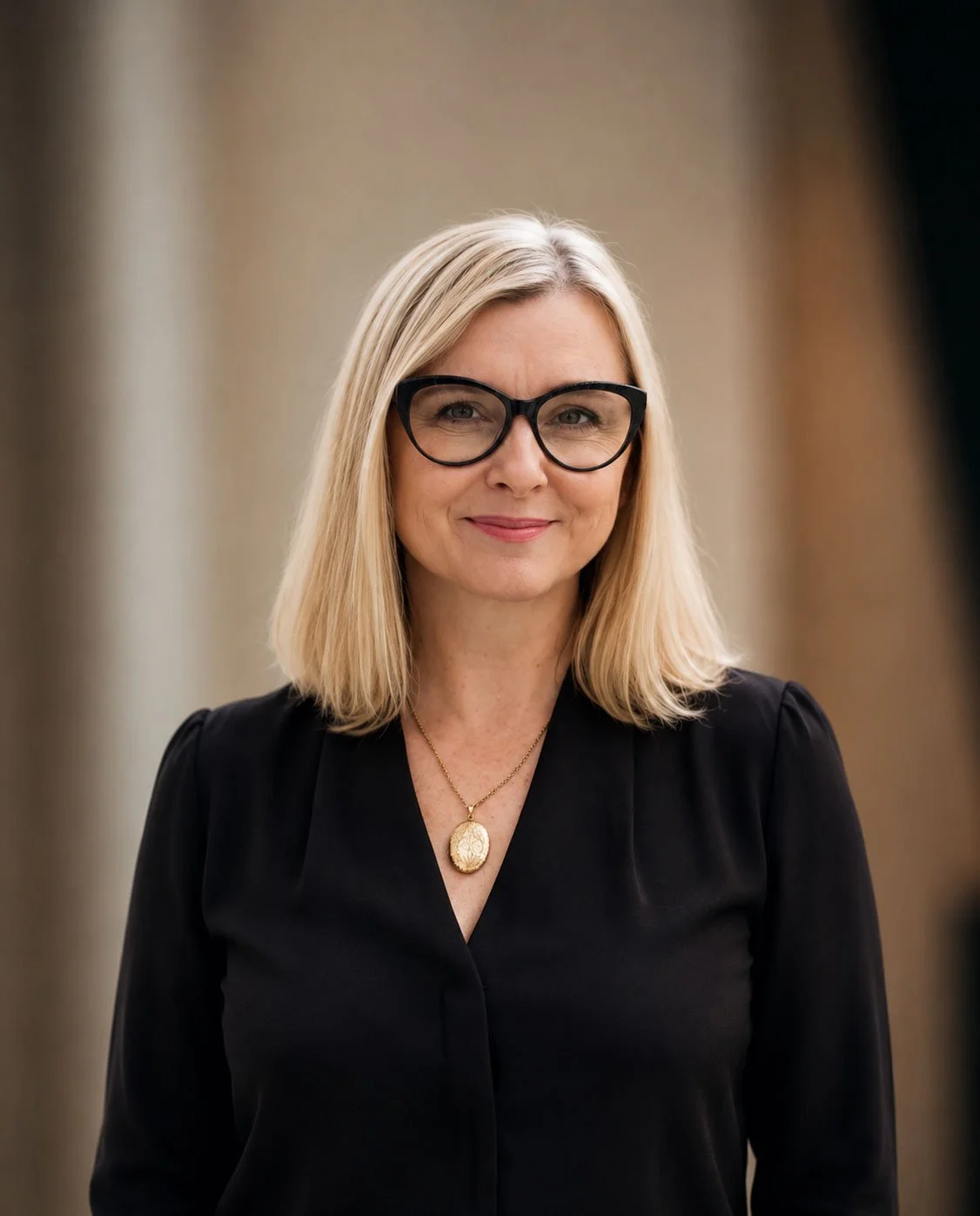 Portrait of a smiling middle-aged woman with blonde hair, black glasses, and wearing a black top with a gold necklace.