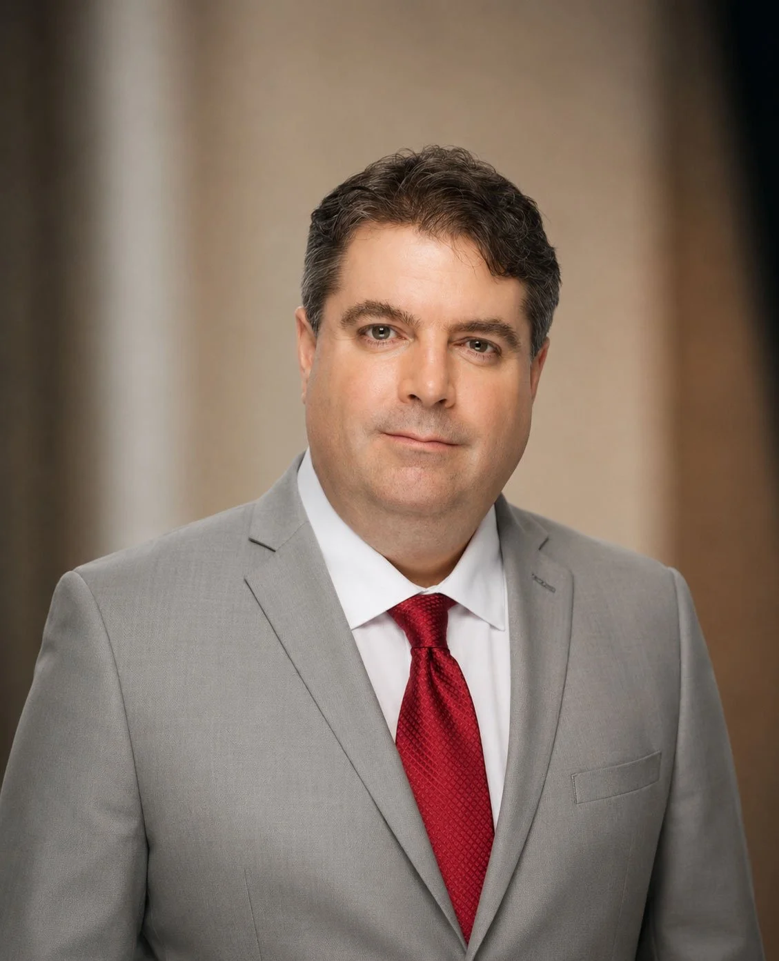 A man with dark hair, dressed in a light gray suit, white shirt, and red tie, posing for a professional portrait with a neutral background.