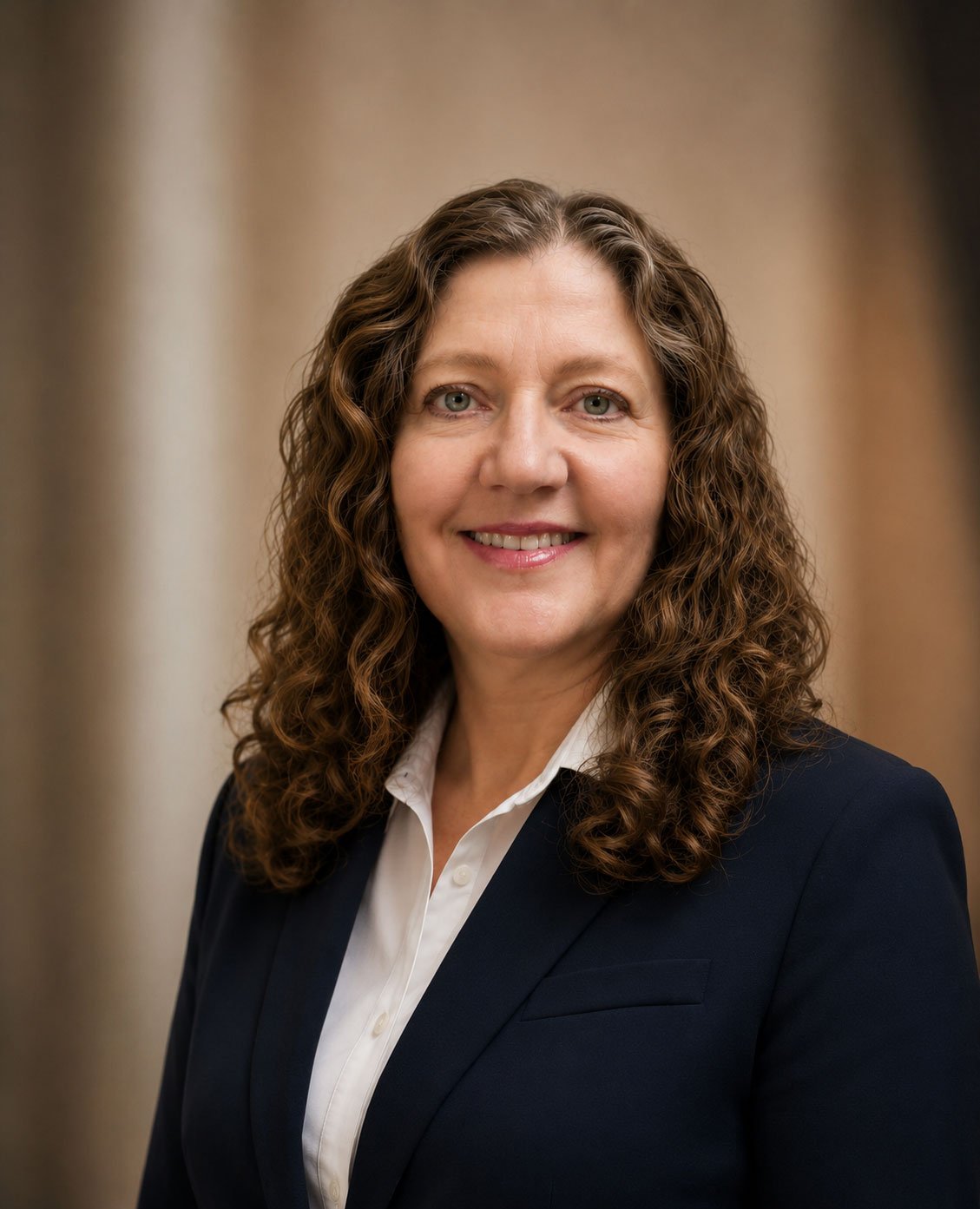 A woman with curly brown hair, wearing a white shirt and navy blazer, smiling in front of a neutral background.