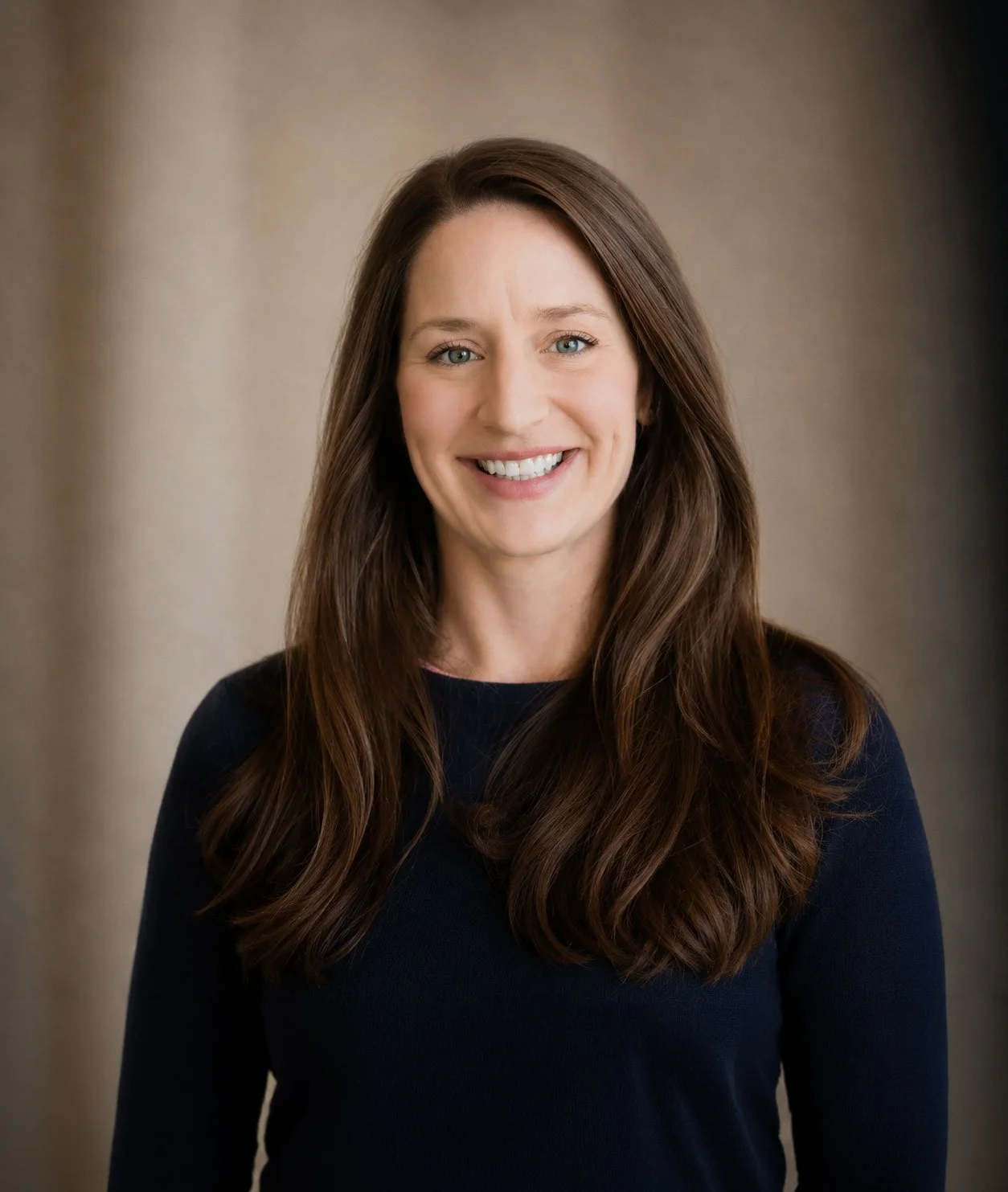 A woman with long brown hair and blue eyes, smiling, wearing a dark long-sleeve top, standing in front of a plain beige background.