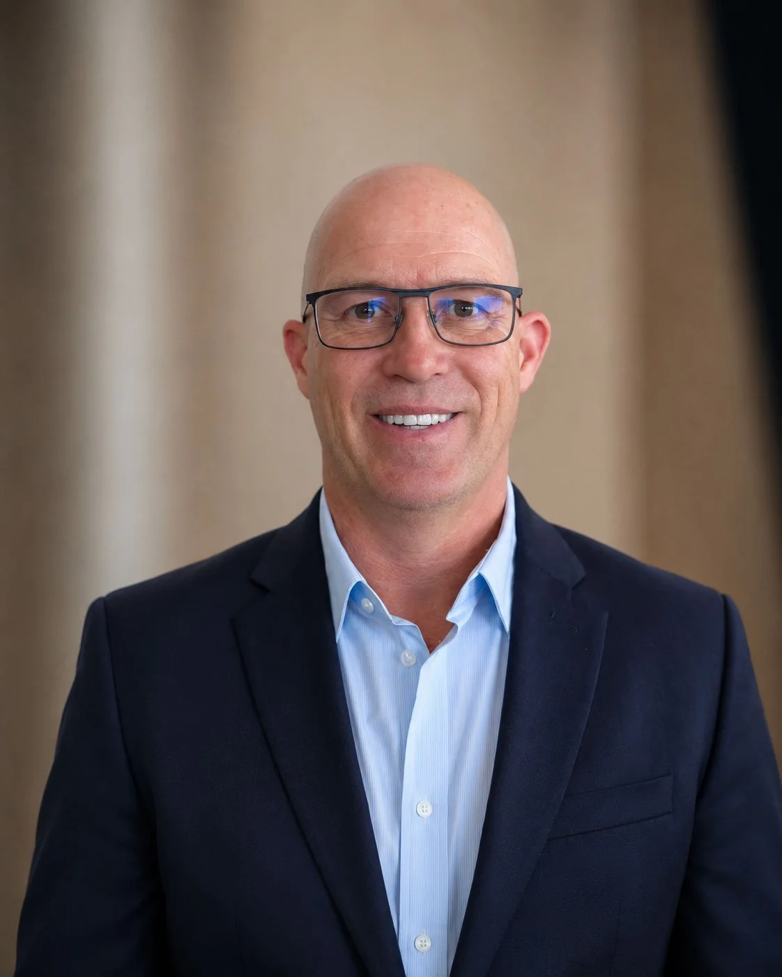 A middle-aged man with a bald head, wearing glasses, a light blue collared shirt, and a navy blazer, smiling at the camera.