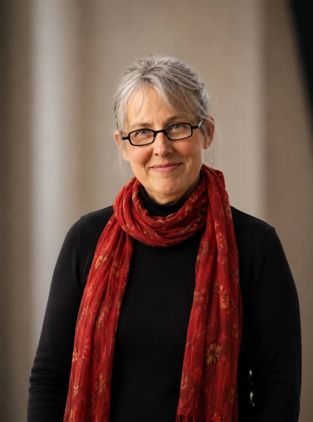 A woman with short gray hair, wearing black glasses, a black top, and a red patterned scarf, smiling at the camera.