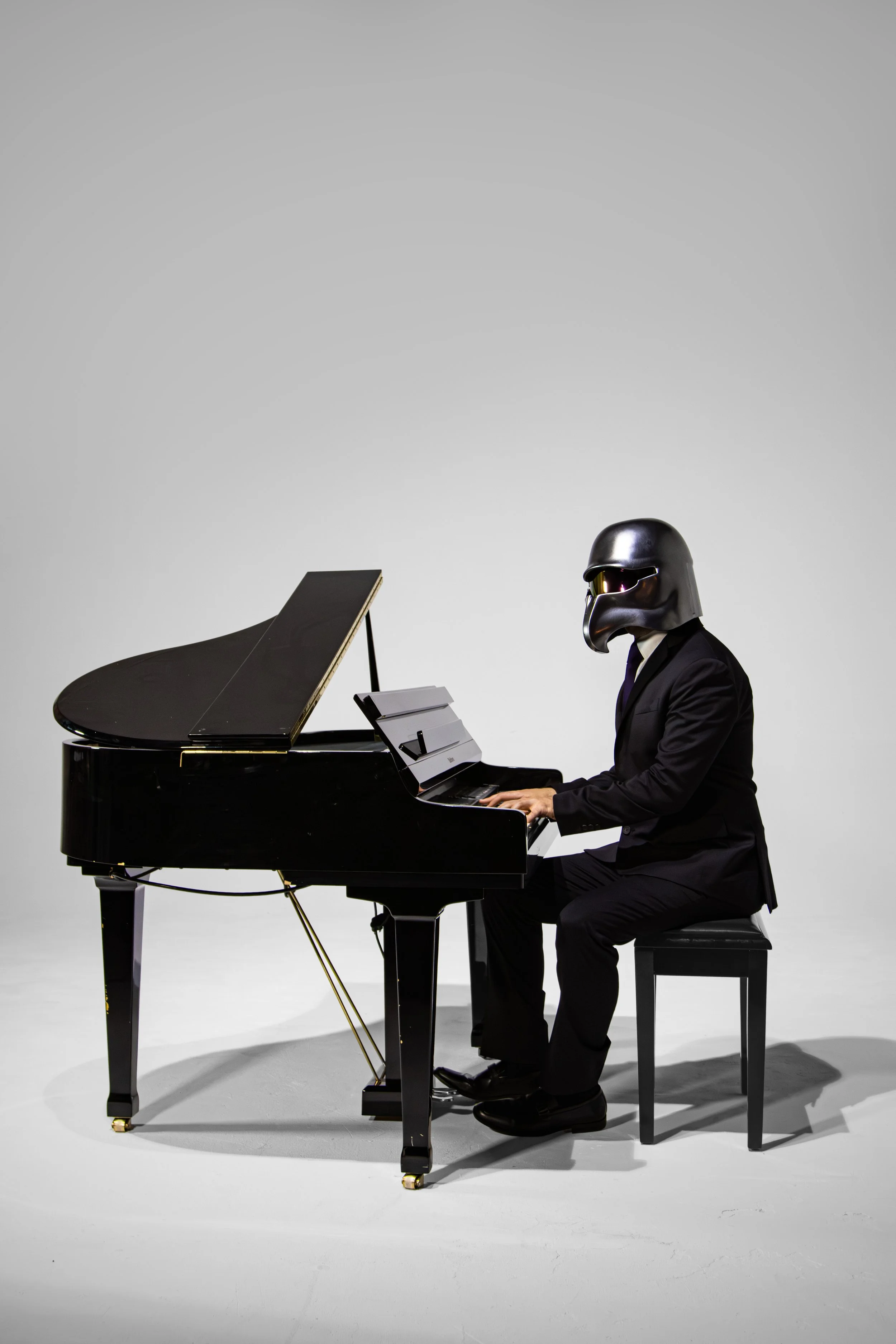 Person wearing a Darth Vader helmet, dressed in a black suit, playing a grand piano in a minimalist setting.